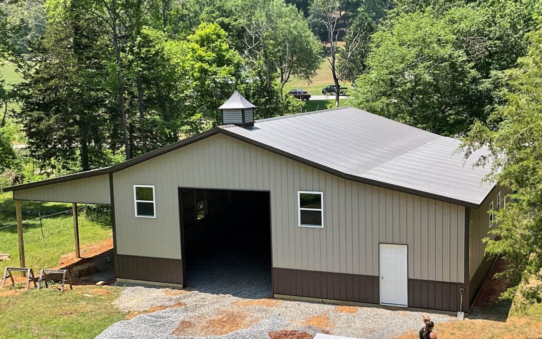 Amish Ridge Metals Mt Perry OH A large, beige metal barn with a gray roof and cupola sits on a gravel driveway surrounded by trees and greenery.