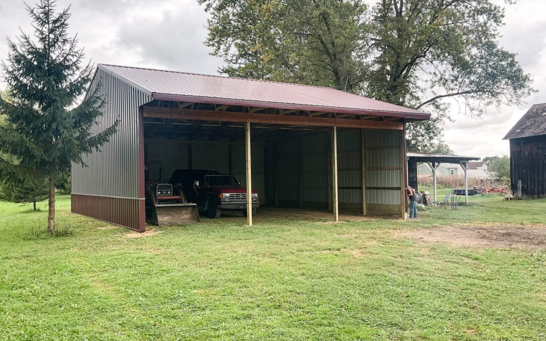 Amish Ridge Metals Mt Perry OH A metal carport with two trucks parked inside stands on a grassy area near trees, with a small shed and swing set visible in the background.
