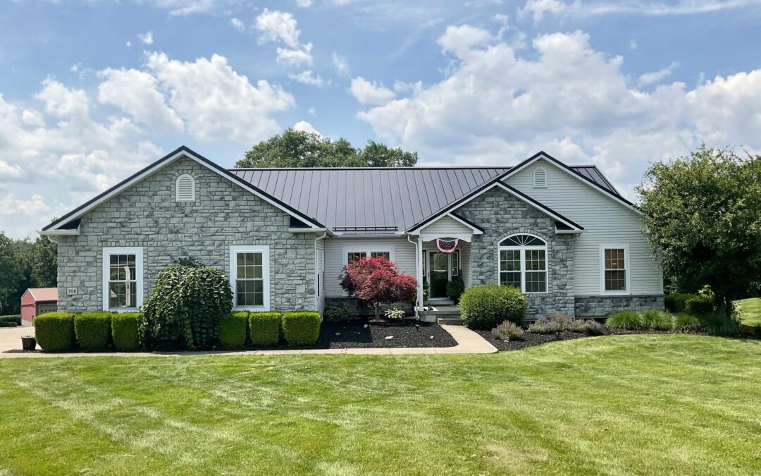 Amish Ridge Metals Mt Perry OH A single-story house with stone and siding exterior, landscaped bushes, arched windows, a black metal roof, and a well-maintained lawn under a partly cloudy sky.