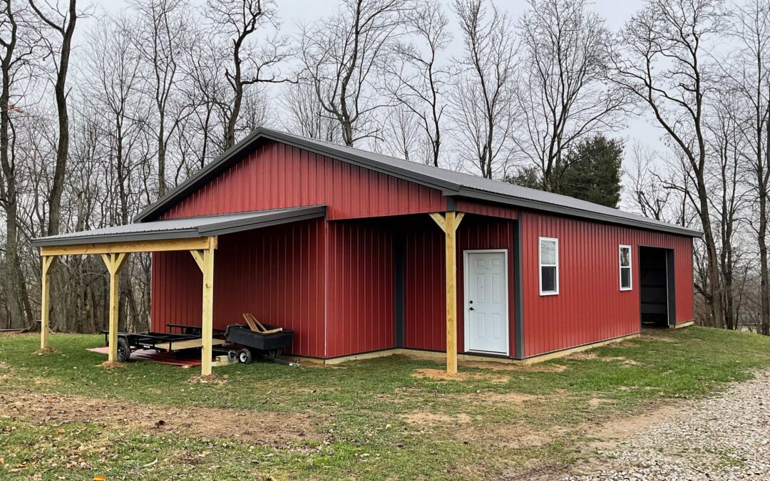Amish Ridge Metals Mt Perry OH A red metal shed with a covered porch and white door stands on a grassy yard, surrounded by leafless trees and a gravel driveway.