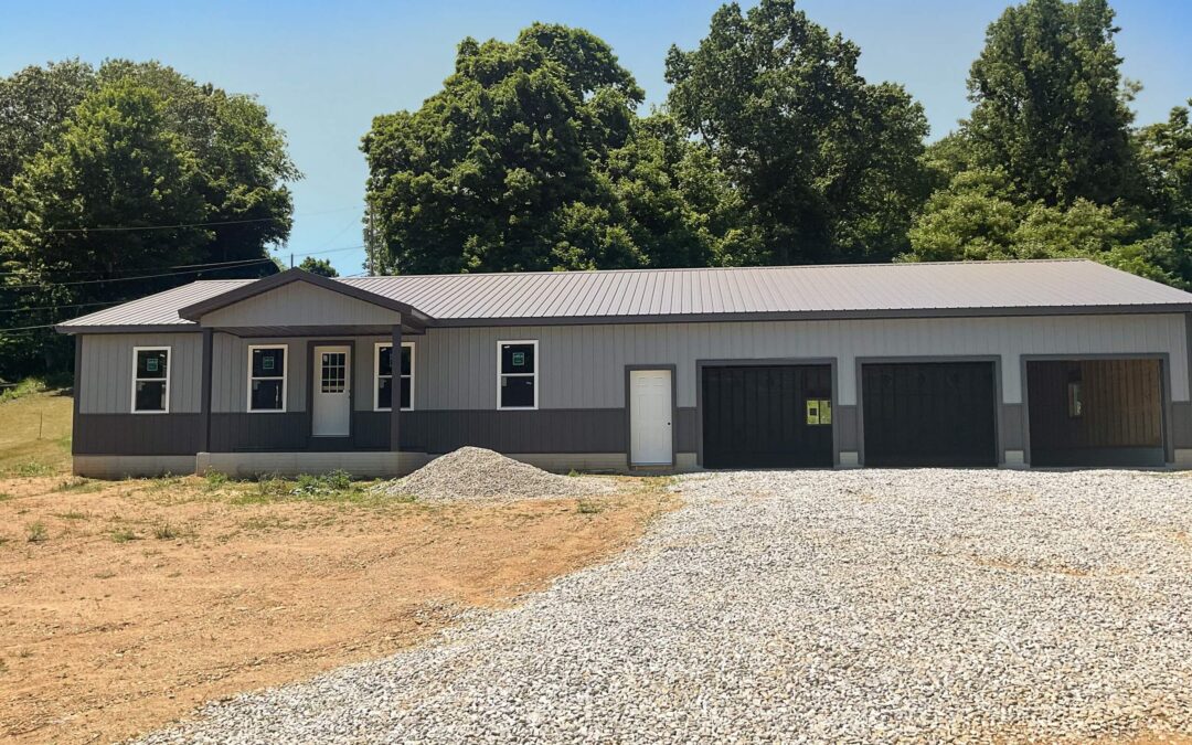 Amish Ridge Metals Mt Perry OH A gray, single-story building with a metal roof, three garage doors, a white entry door, and gravel driveway, set against a backdrop of trees and blue sky.