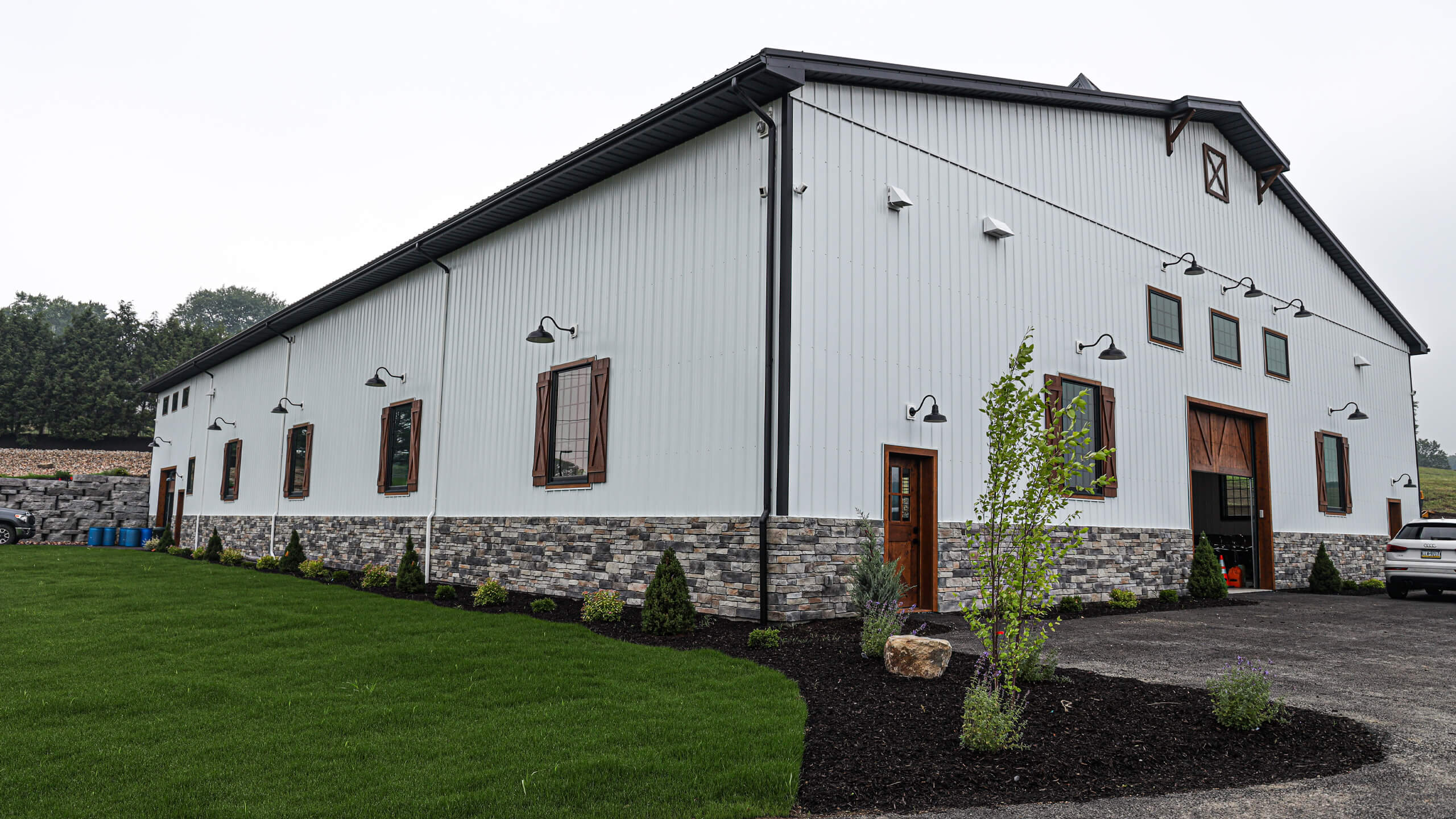 3.4.26 Dave's Metal Roofing Gallery 3 A large white barn-style building with a stone foundation, multiple windows and doors, exterior lights, landscaped mulch beds, and a paved driveway on a green lawn.