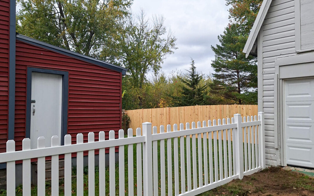 Advanced Privacy Fencing Ovid NY White picket fence separates a red building and a white building with a garage door; trees and a wooden fence are in the background under a cloudy sky.