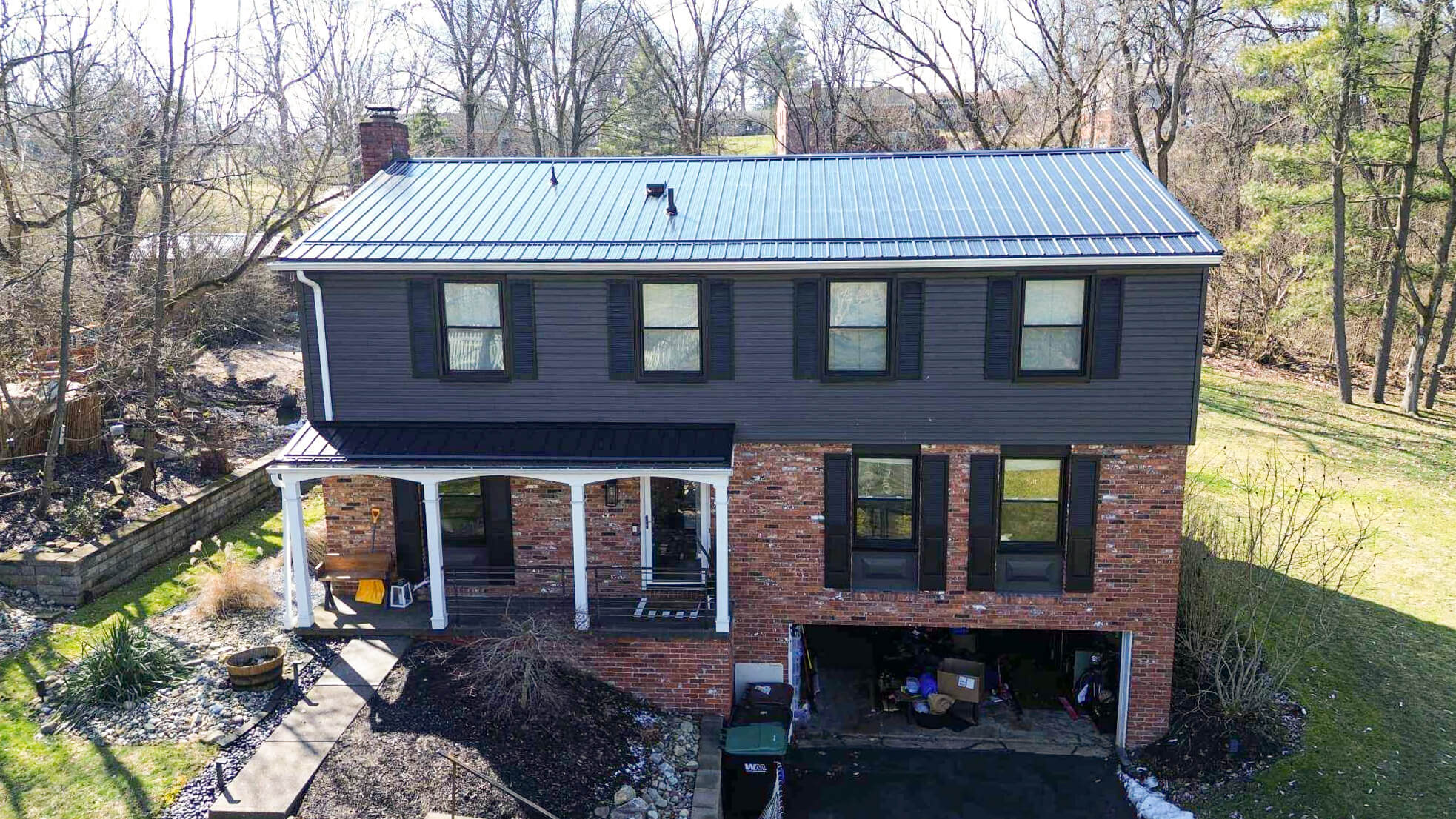Two-story brick and dark siding house with a metal roof, front porch, black shutters, and an open garage, surrounded by leafless trees and a grassy yard.