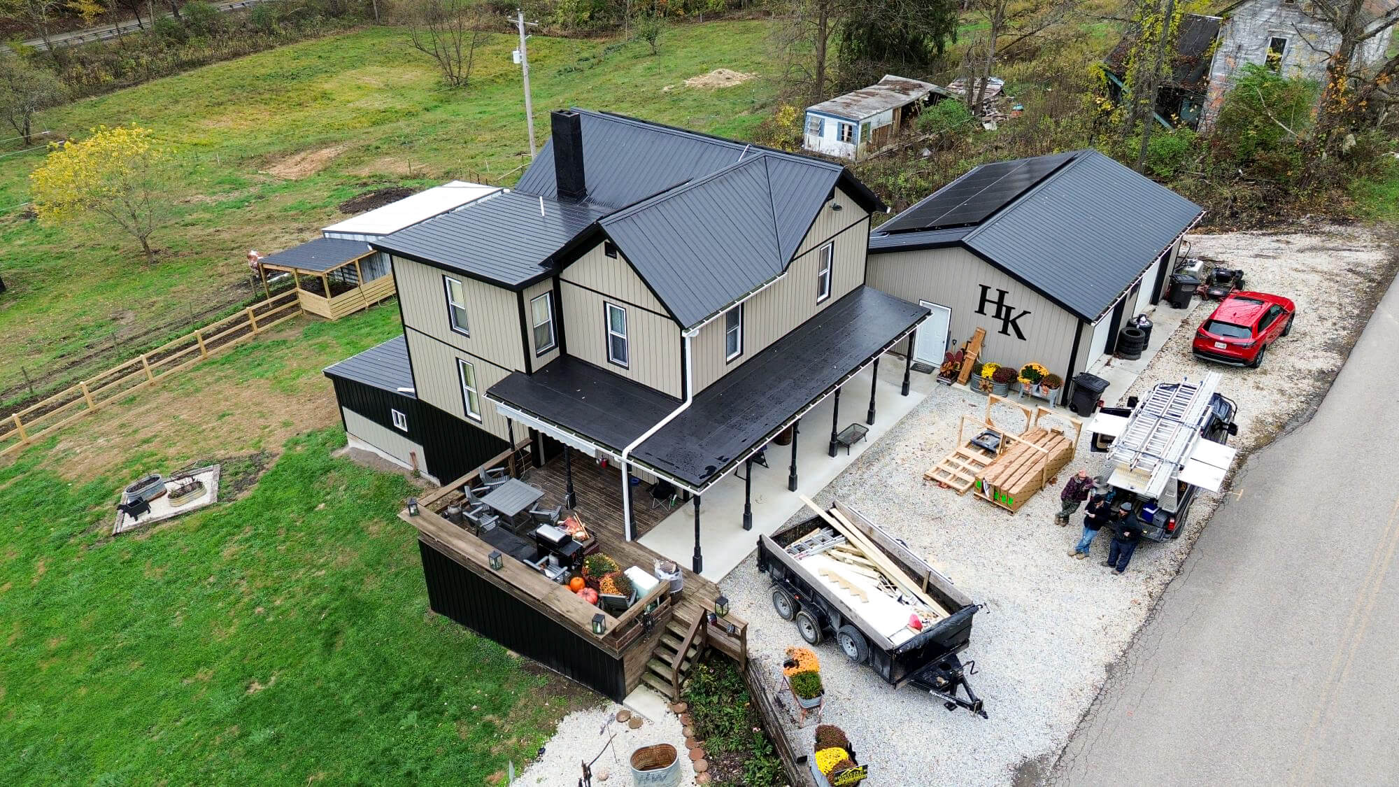 Aerial view of a beige house with a black metal roof, a detached garage, construction materials, vehicles, and people standing on a gravel driveway.