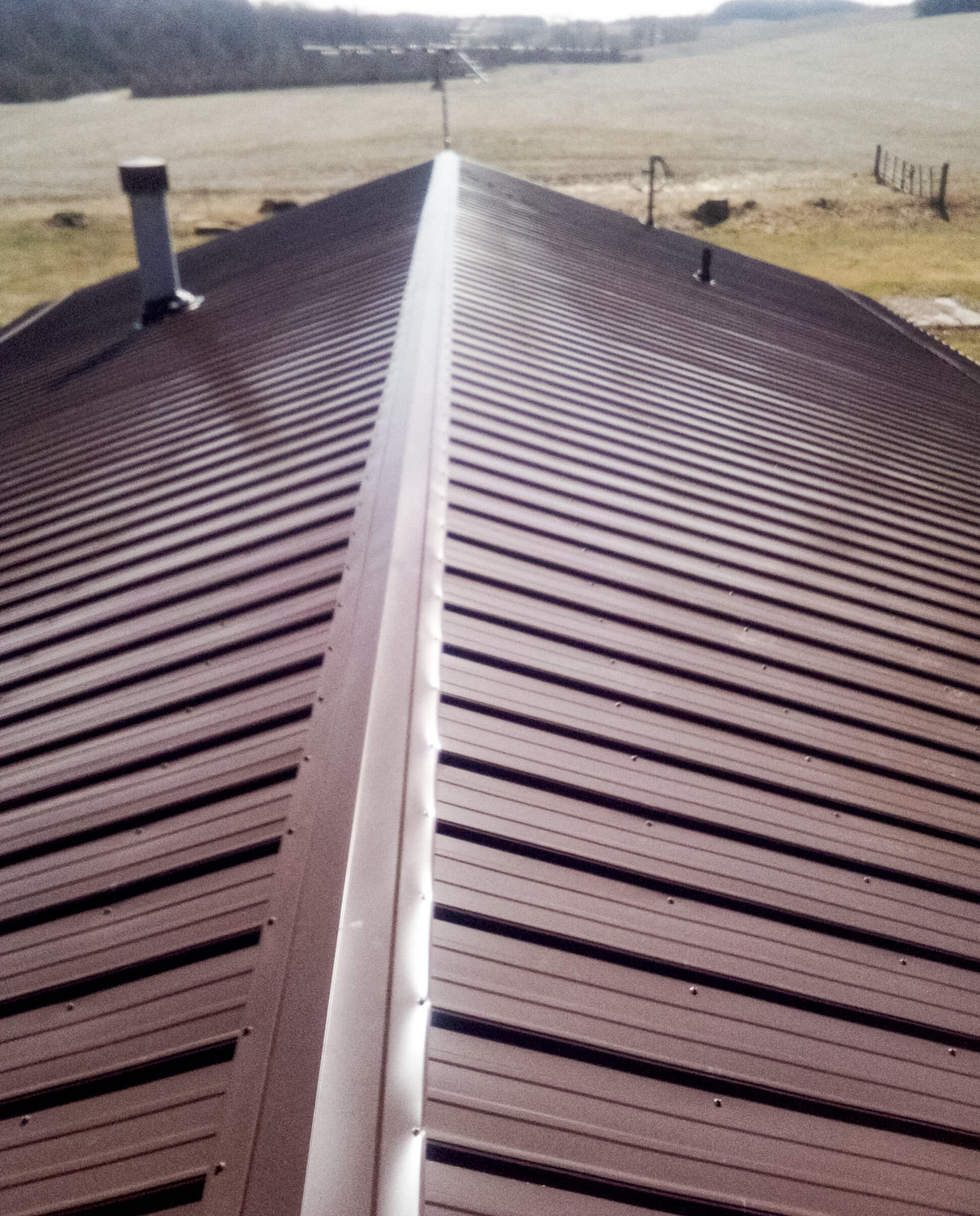 A brown metal roof with a central ridge cap, viewed from above, with a rural landscape and a field in the background.