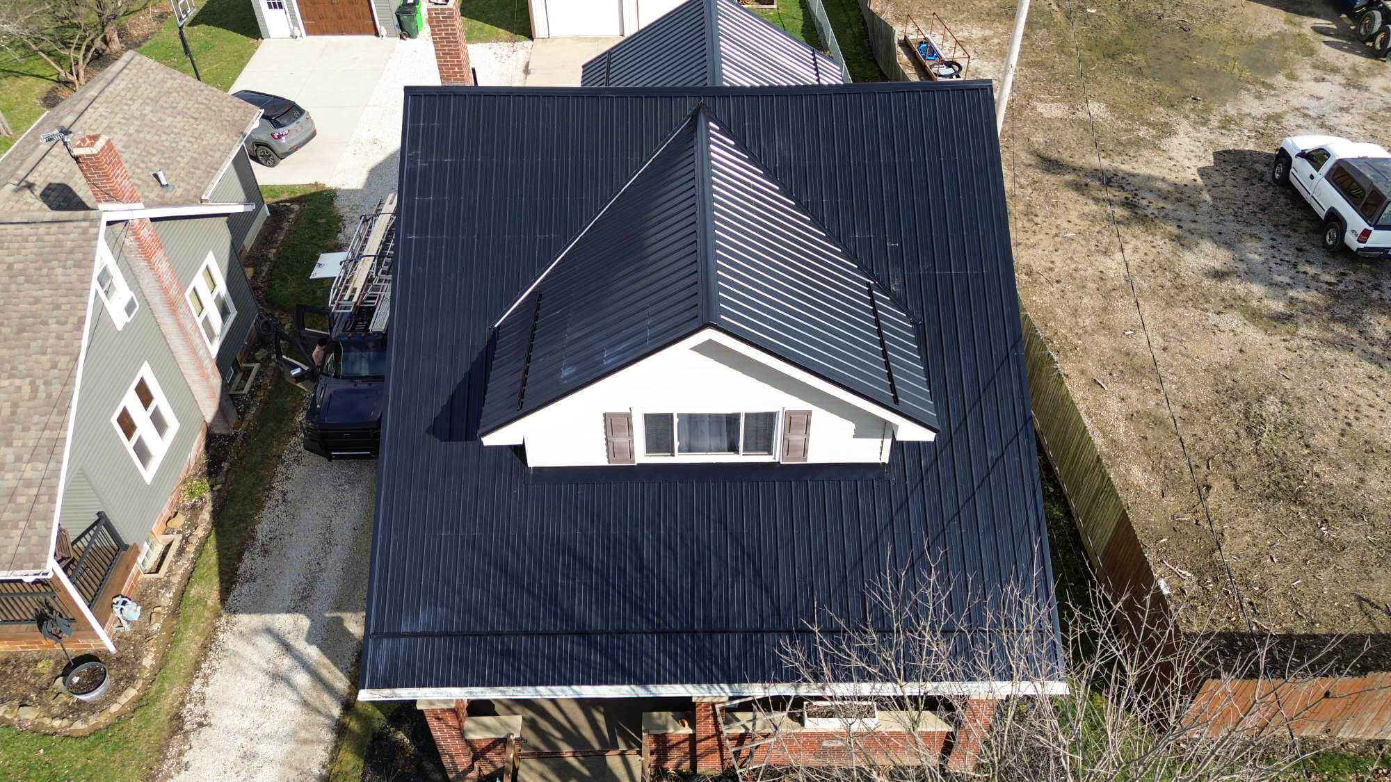 Aerial view of a house with a black metal roof and dormer, surrounded by neighboring homes, driveway, and a car parked nearby.