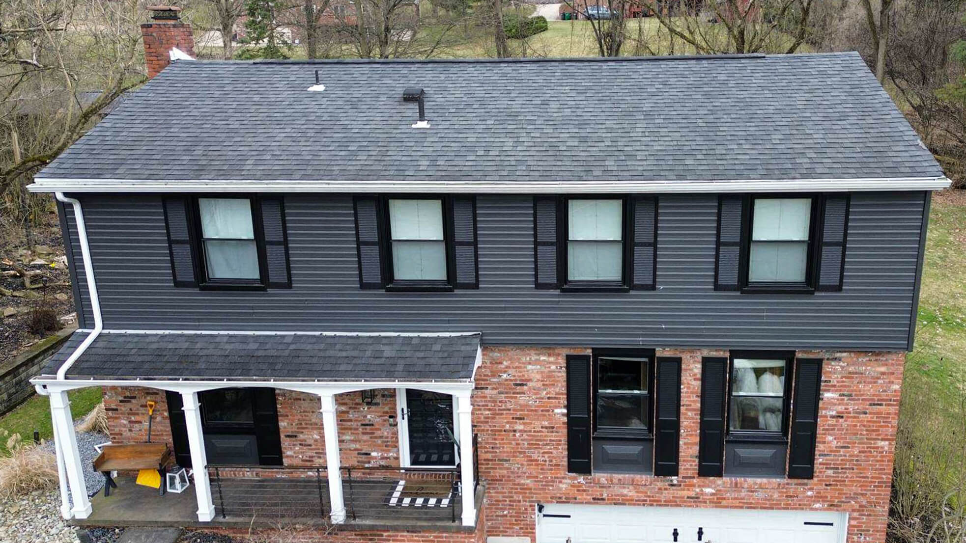 Two-story house with a gray shingle roof, dark gray siding on the upper floor, red brick on the lower level, black shutters, and a white front porch.