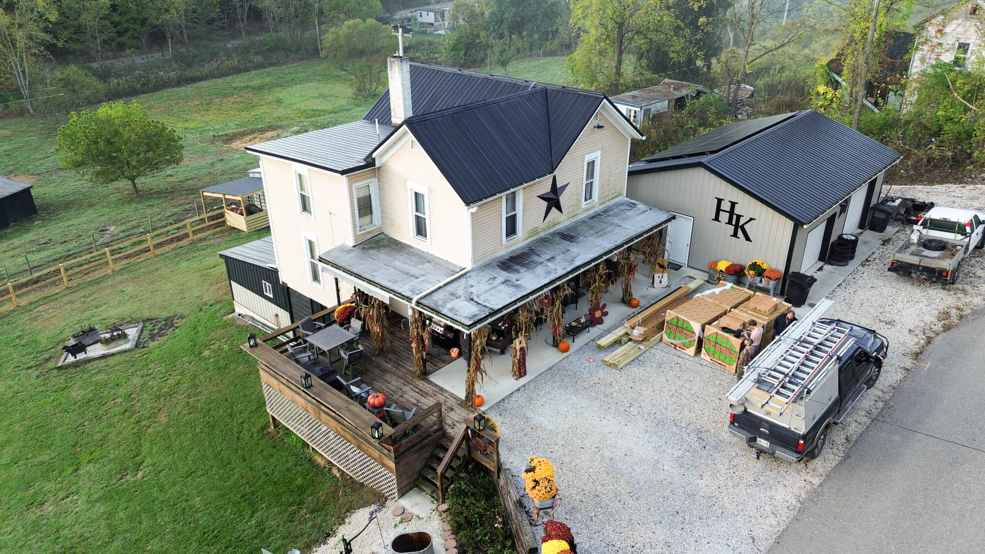 Aerial view of a farmhouse-style store with a front porch decorated for fall, adjacent to a building marked "HK," parked vehicles, and a gravel driveway.
