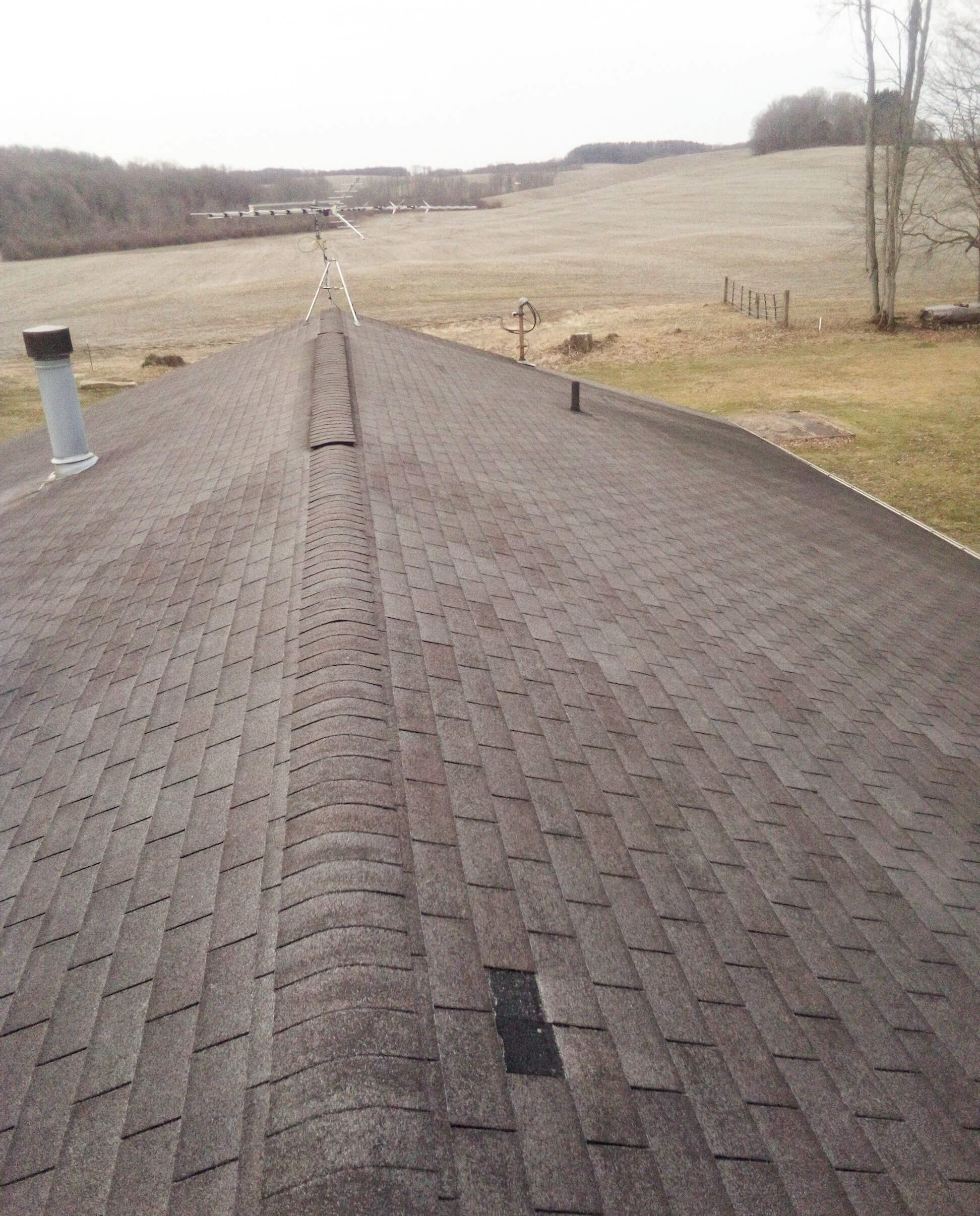 View of a house roof with asphalt shingles; one shingle near the center is missing. Open fields and trees are visible in the background.
