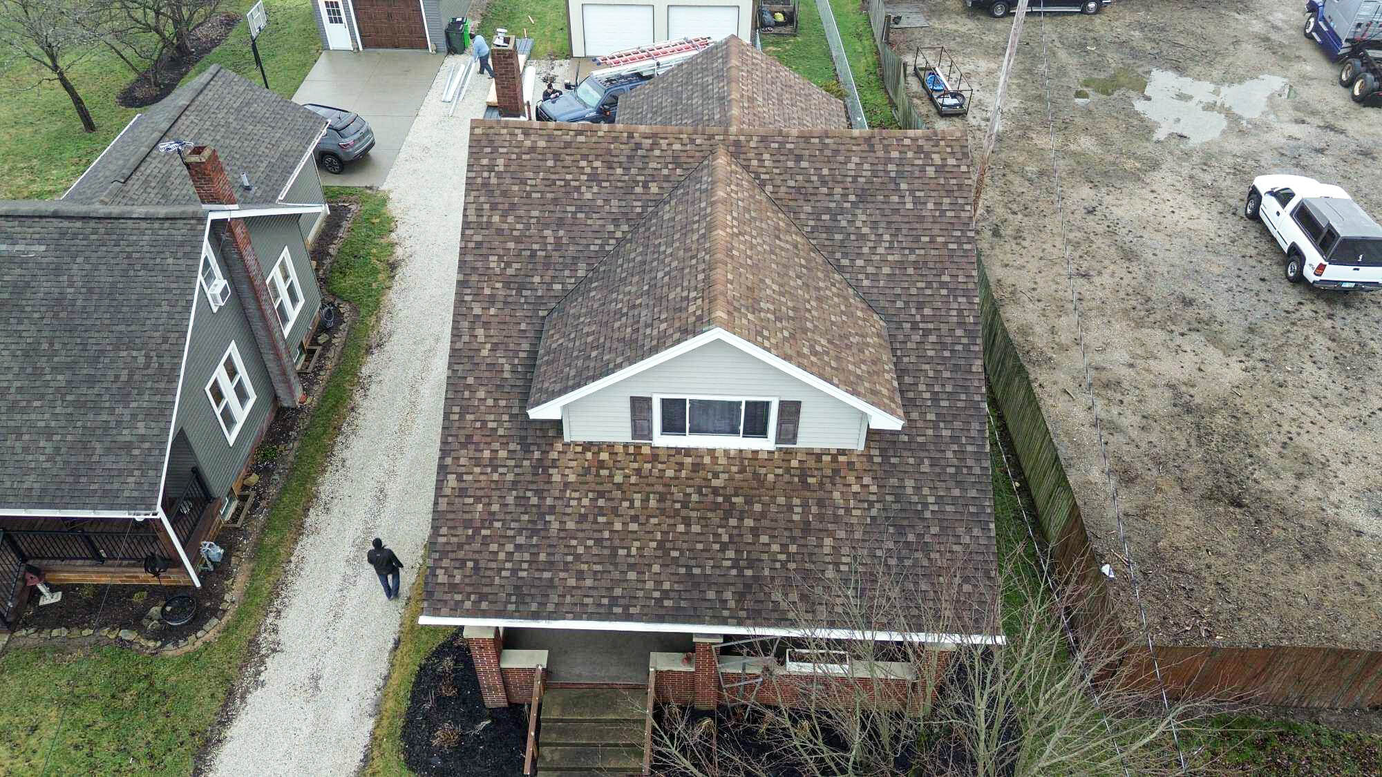 Aerial view of a house with a brown shingle roof, a driveway, and a detached garage. Neighboring houses and vehicles are visible nearby.