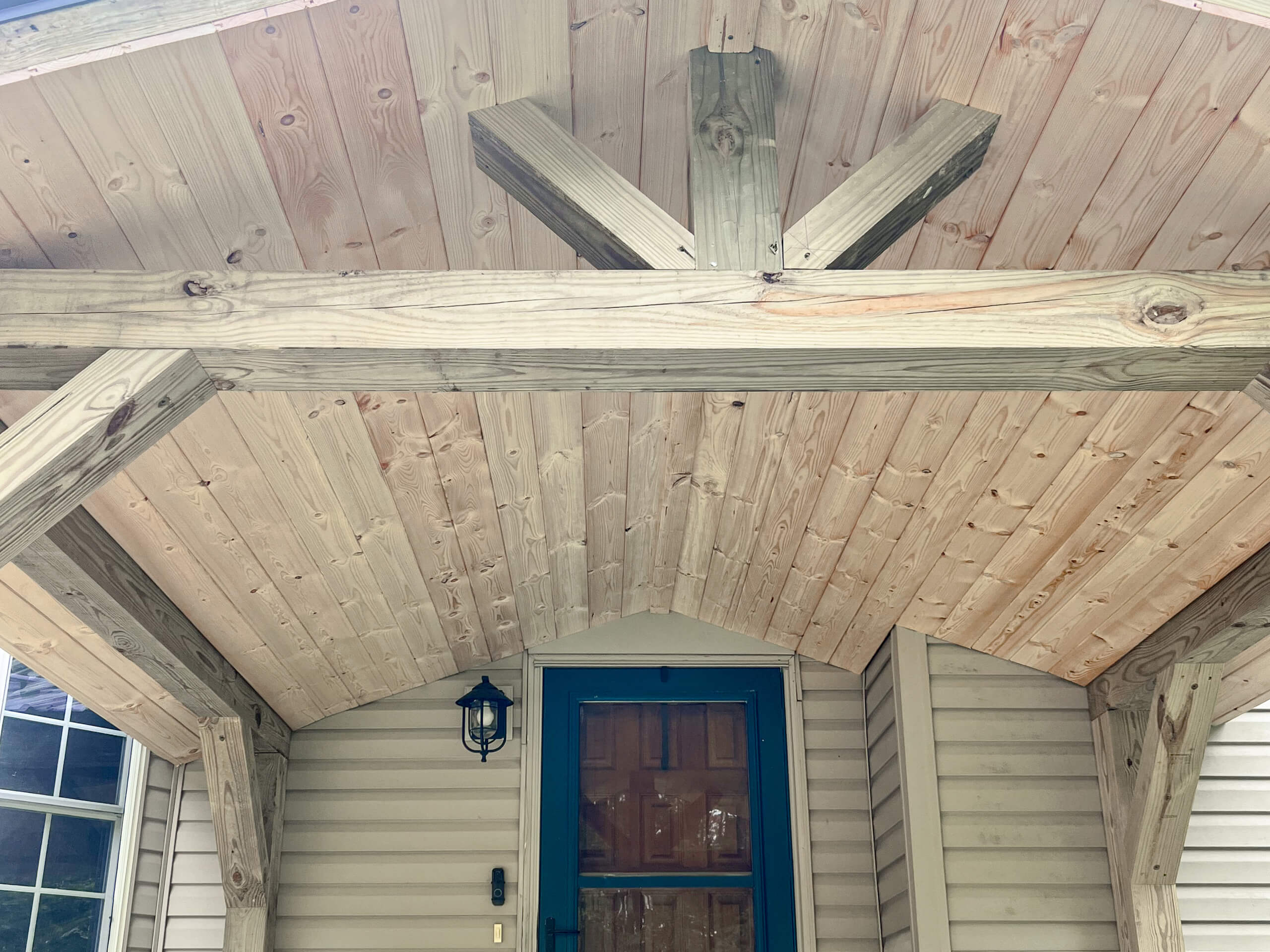 Crawford County Roofing Spartansburg PA Wooden porch roof structure with exposed beams above a door, next to a window on a house with beige siding. A black outdoor light fixture is mounted next to the door.