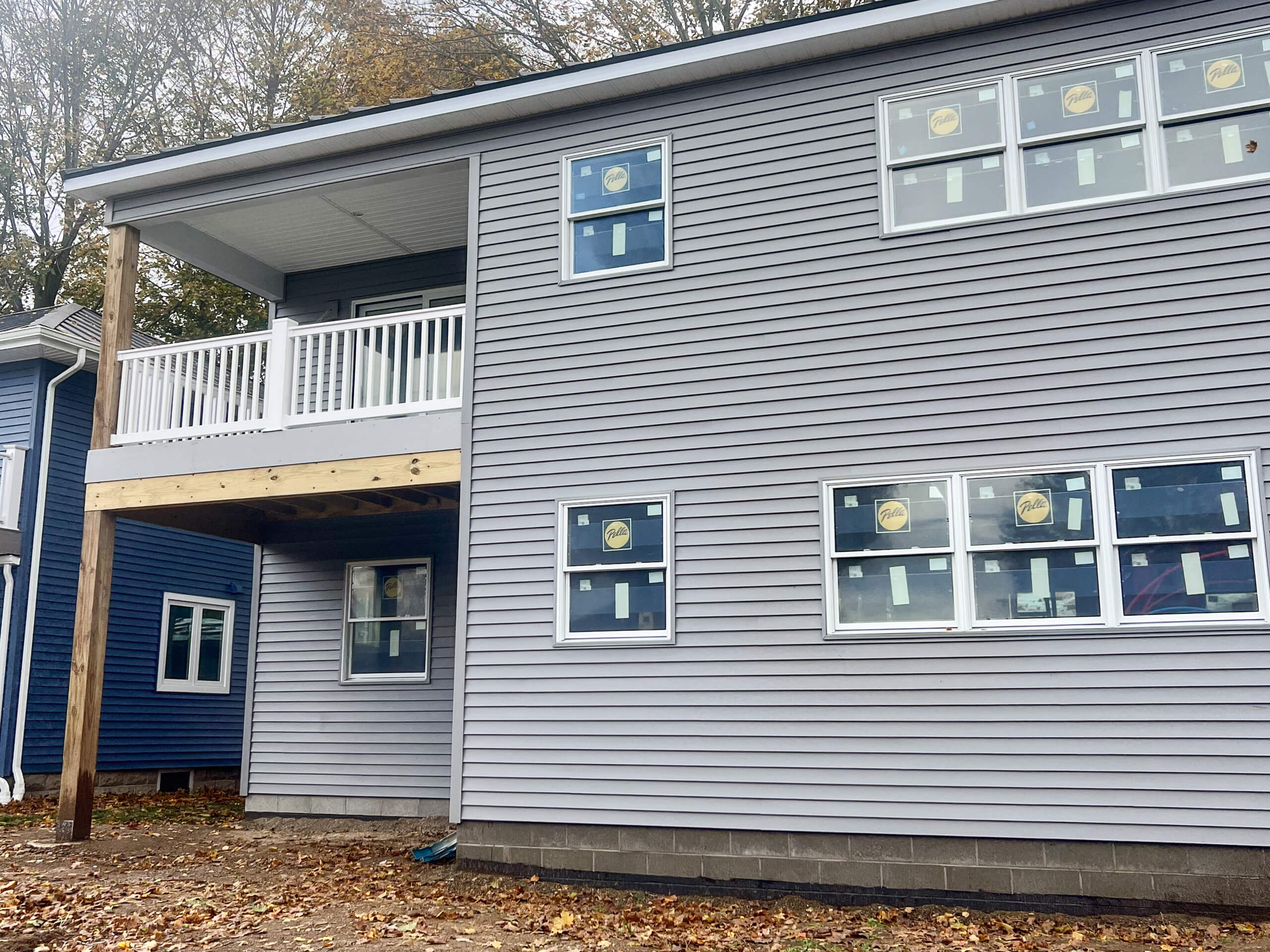 Crawford County Roofing Spartansburg PA Two-story house with gray siding, multiple windows covered with labels, a second-floor balcony, and unfinished ground in front.