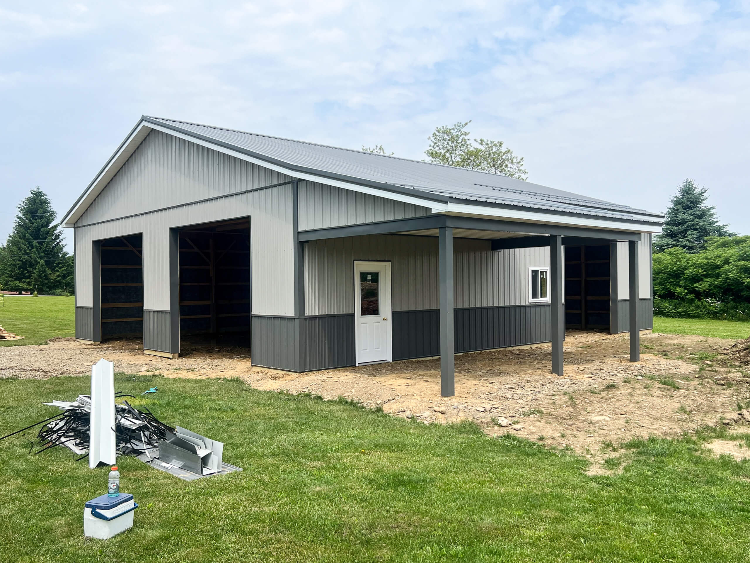 Crawford County Roofing Spartansburg PA A newly constructed gray metal barn with open garage bays and a covered entry door, situated on a grassy yard with construction materials nearby.