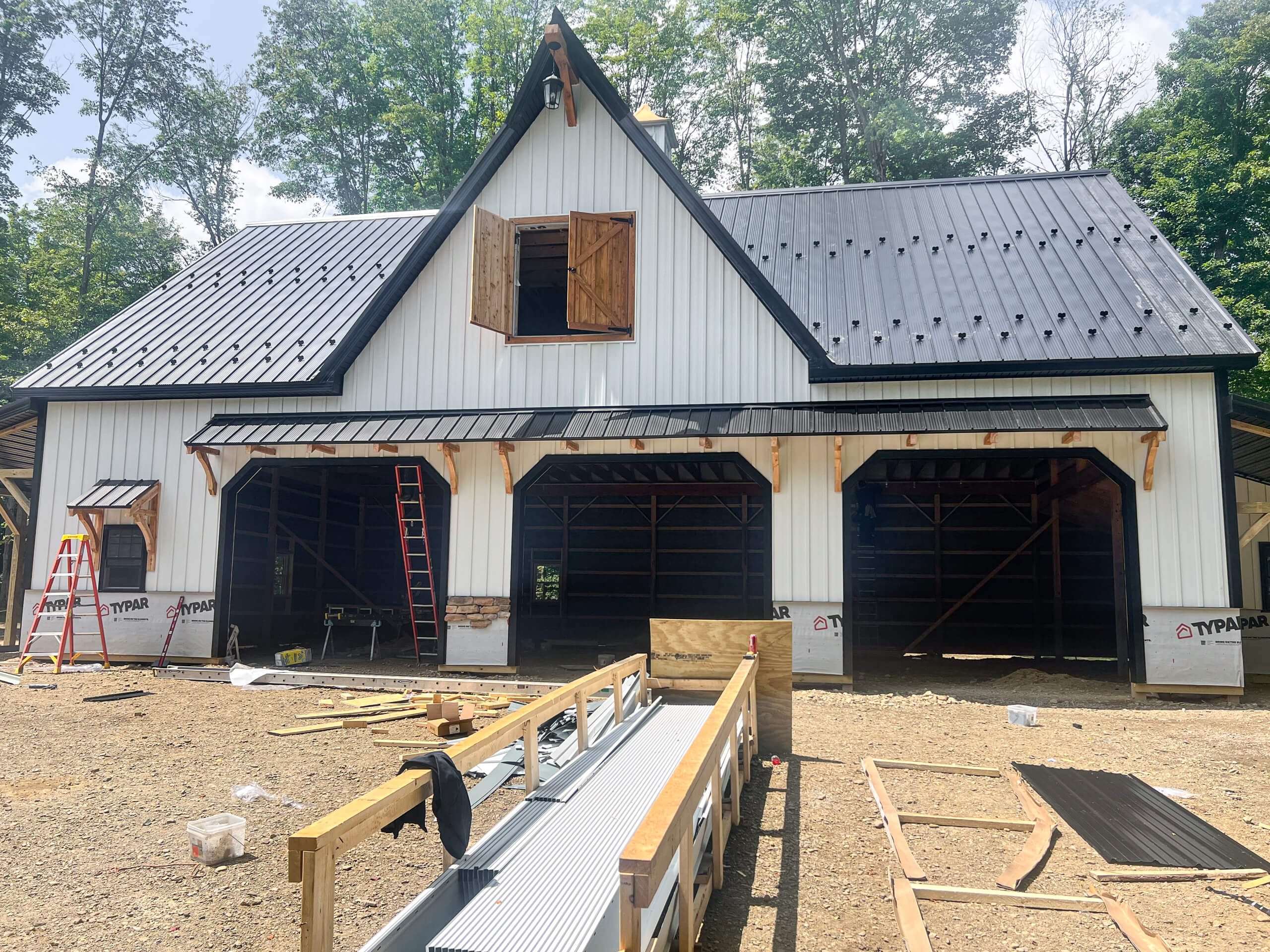 Crawford County Roofing Spartansburg PA A partially constructed building with three garage door openings, black metal roof, ladders, and construction materials scattered on the ground. Trees are visible in the background.