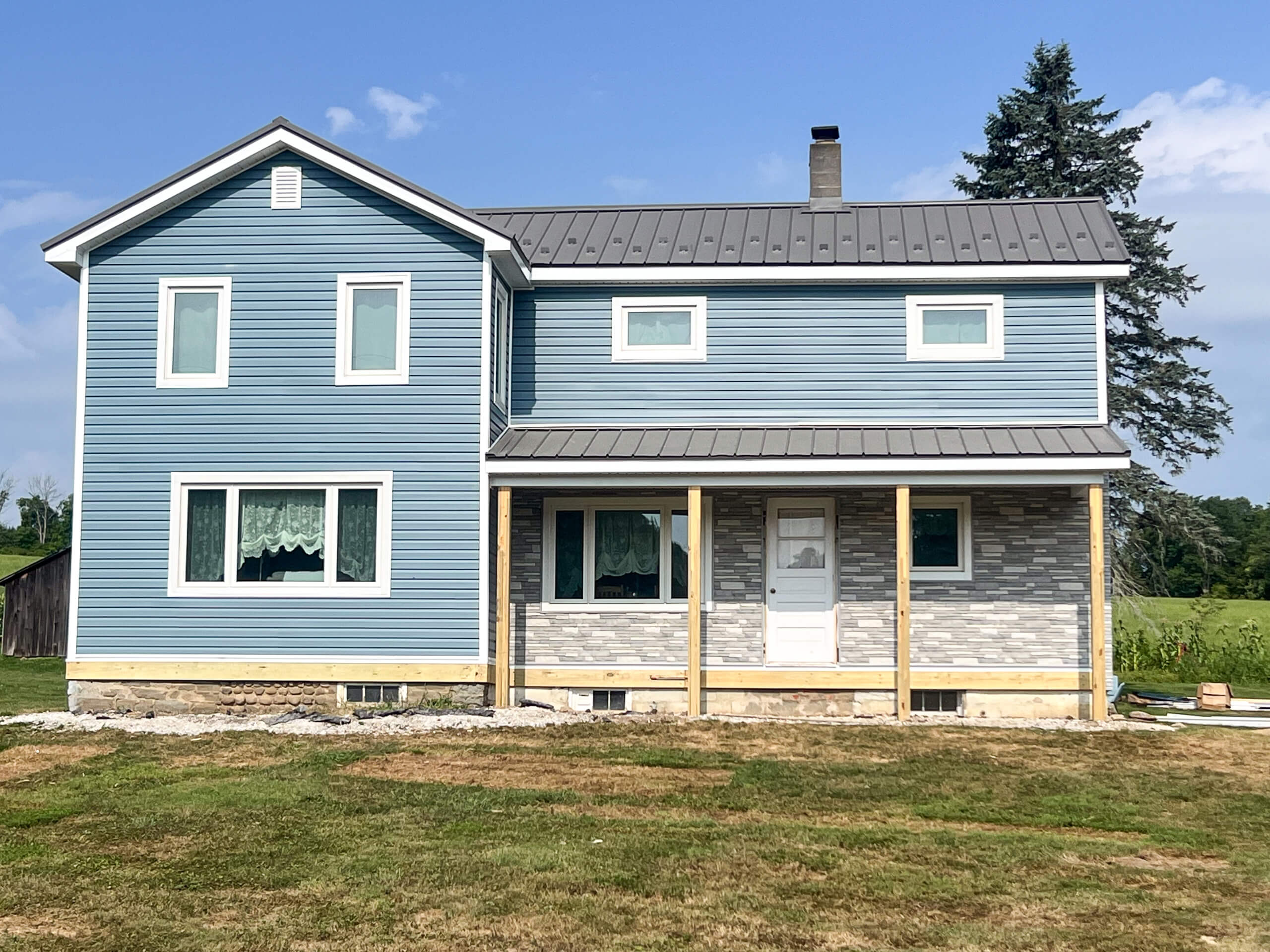 Crawford County Roofing Spartansburg PA A two-story blue house with white trim, a metal roof, and a partially finished porch stands on a grassy lot under a clear sky.