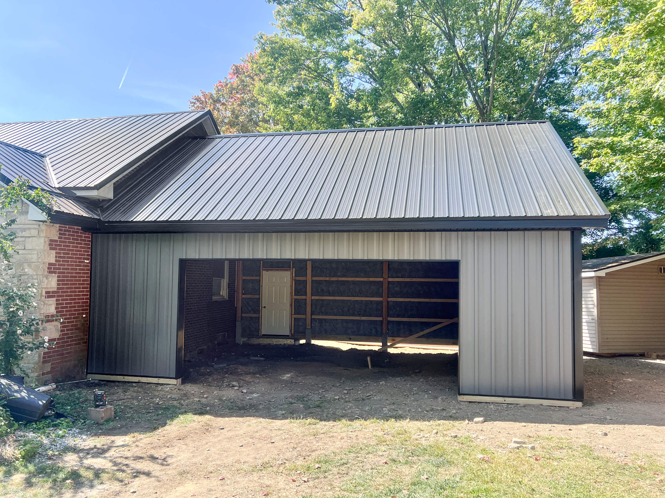 Crawford County Roofing Spartansburg PA A partially constructed garage with a metal roof and siding, open at the front, showing exposed framing and a door at the back, surrounded by trees and dirt ground.