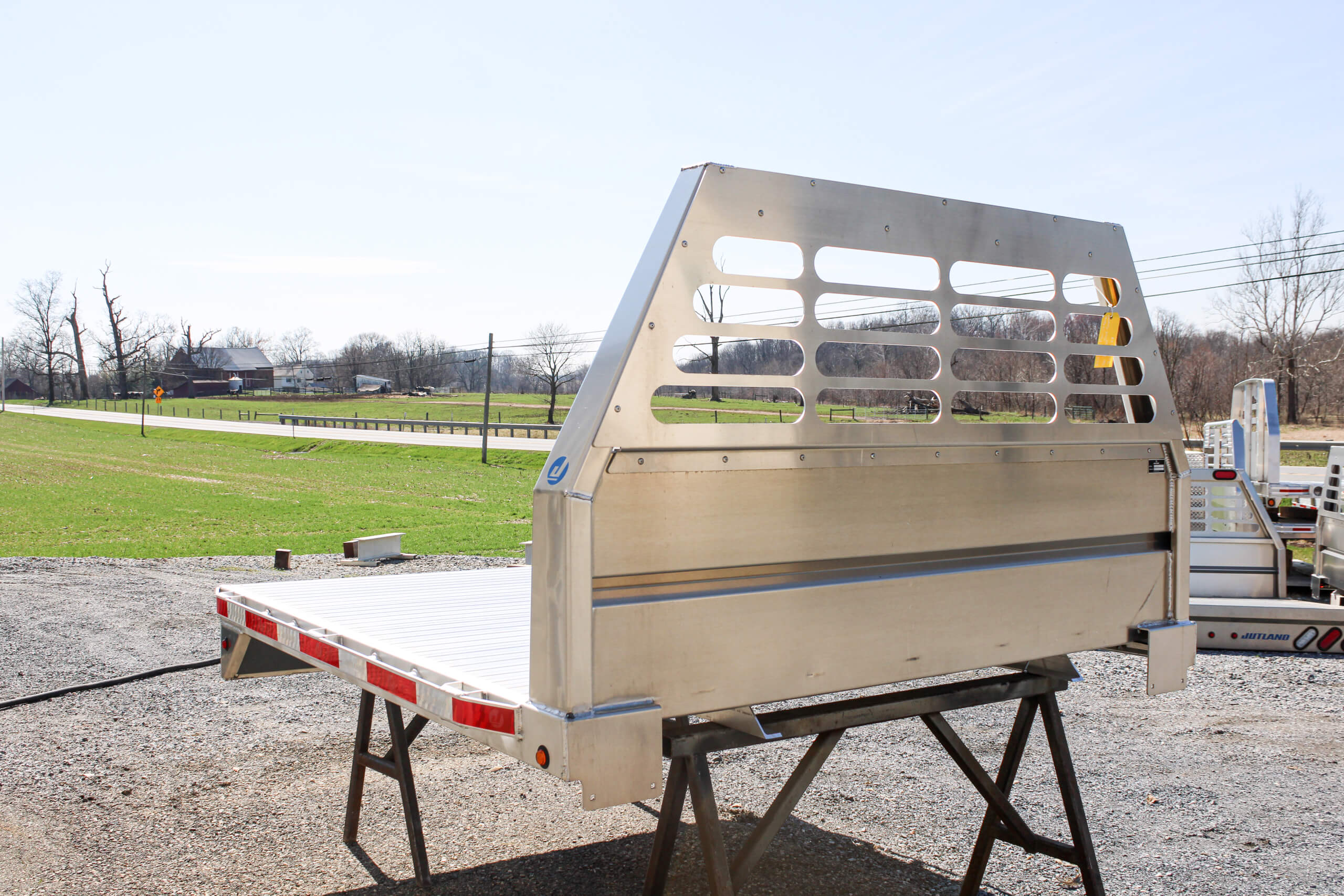 Flatbed aluminum truck bed displayed on metal stands outdoors near a grassy field and road.