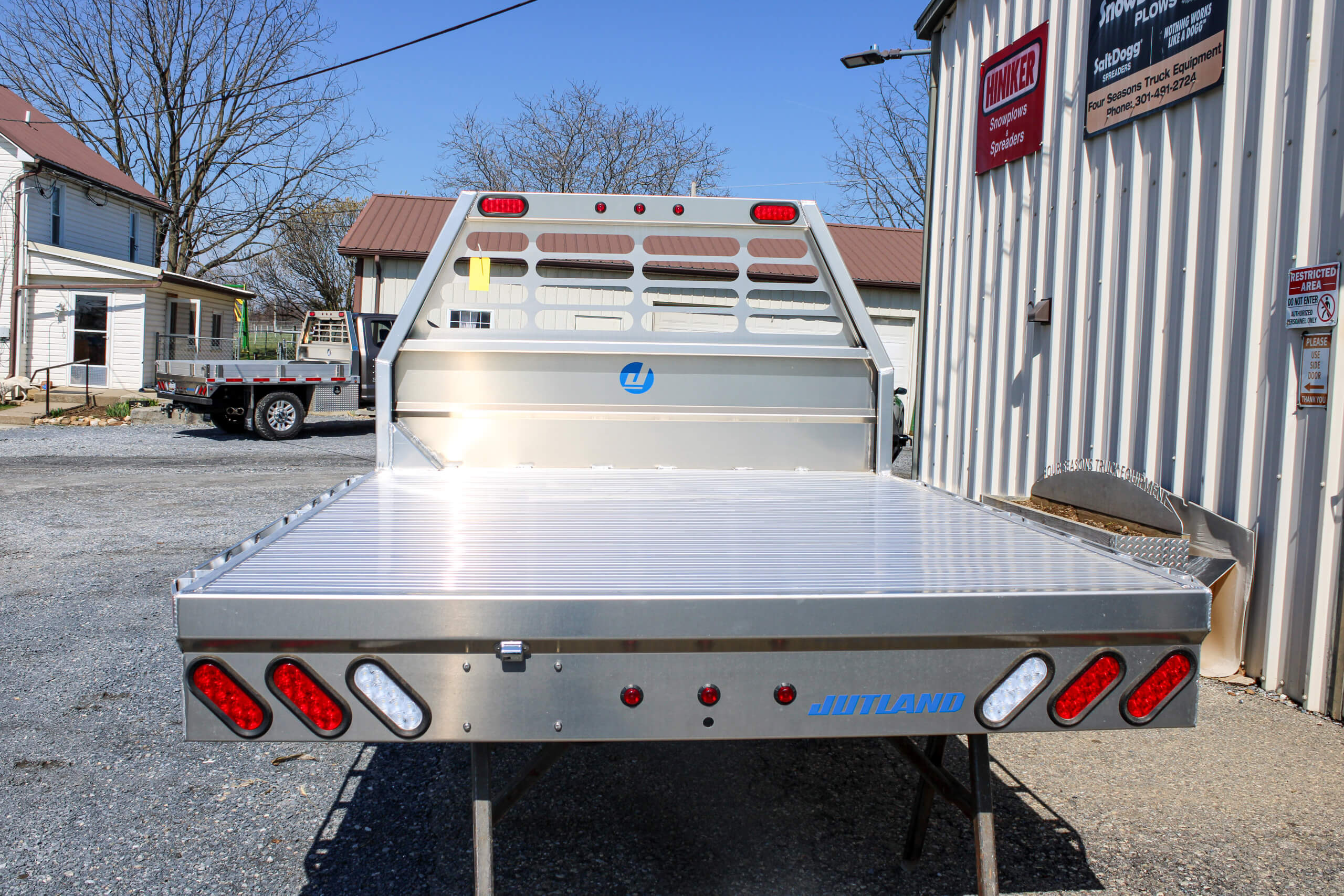 An aluminum flatbed truck body with red and white lights is parked outside a metal building on a gravel lot.