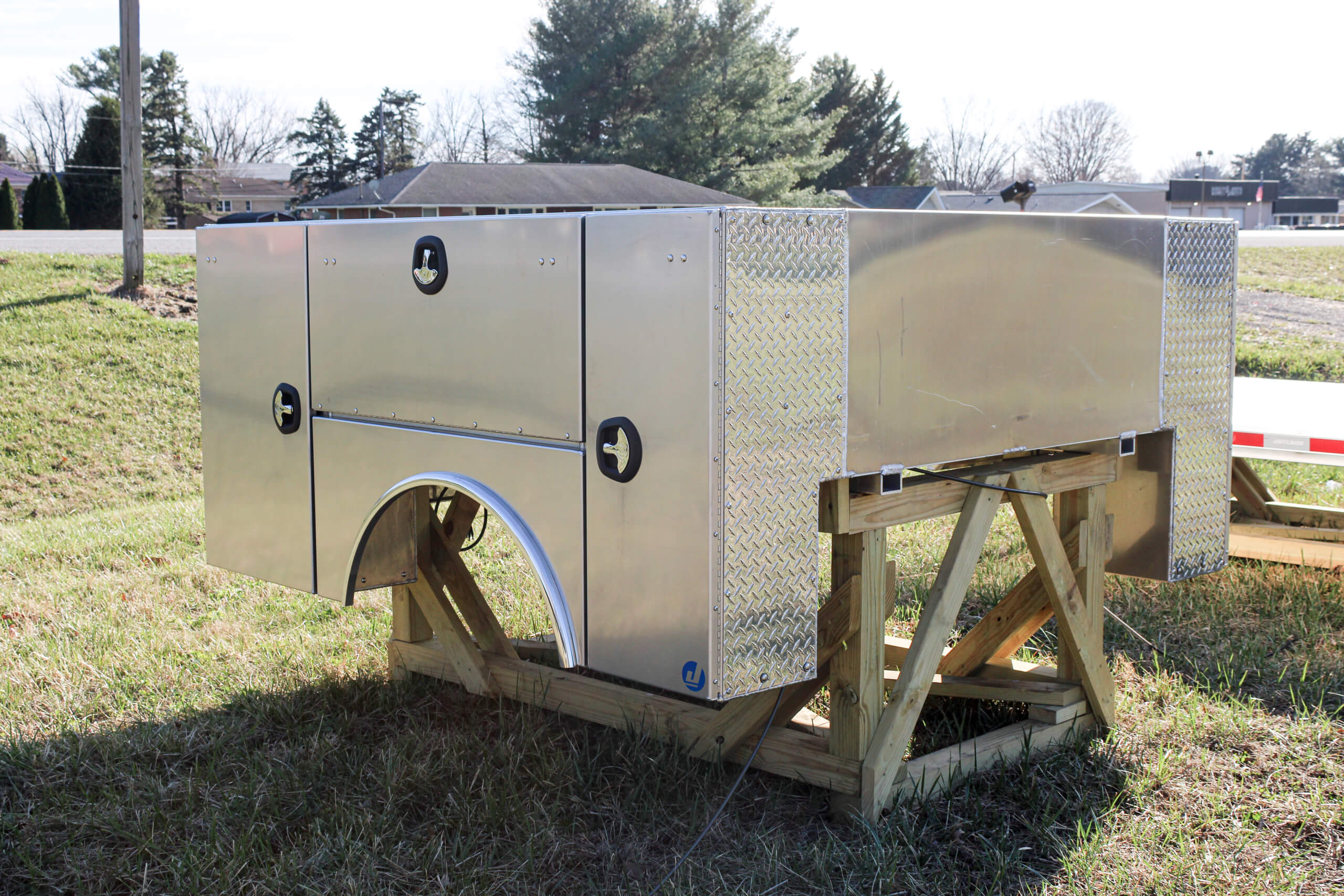 Metal utility truck bed mounted on a wooden frame outdoors, with grass and trees in the background.