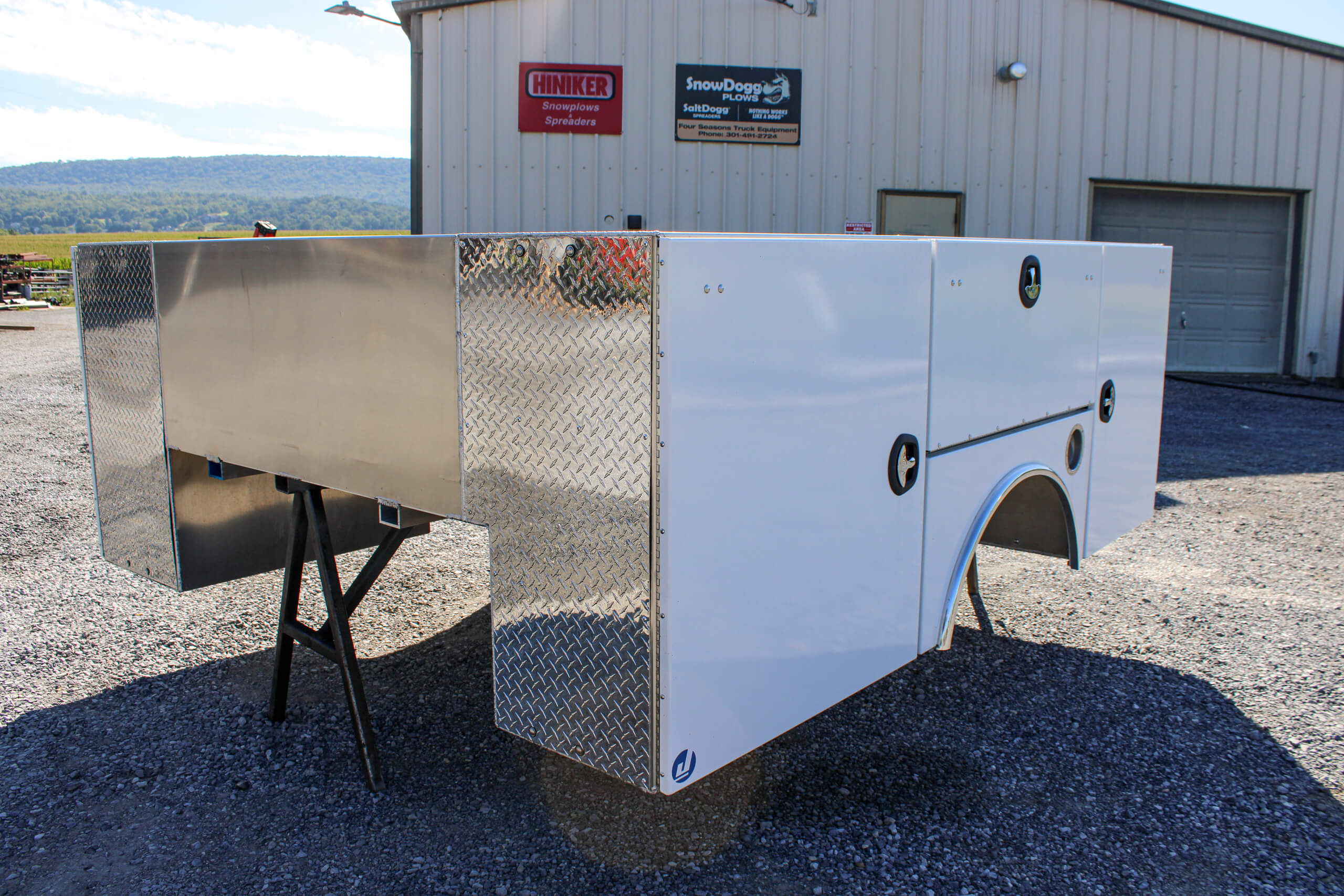 White utility truck bed with diamond plate accents displayed outdoors on stands in a gravel lot, with a metal building and signage in the background.