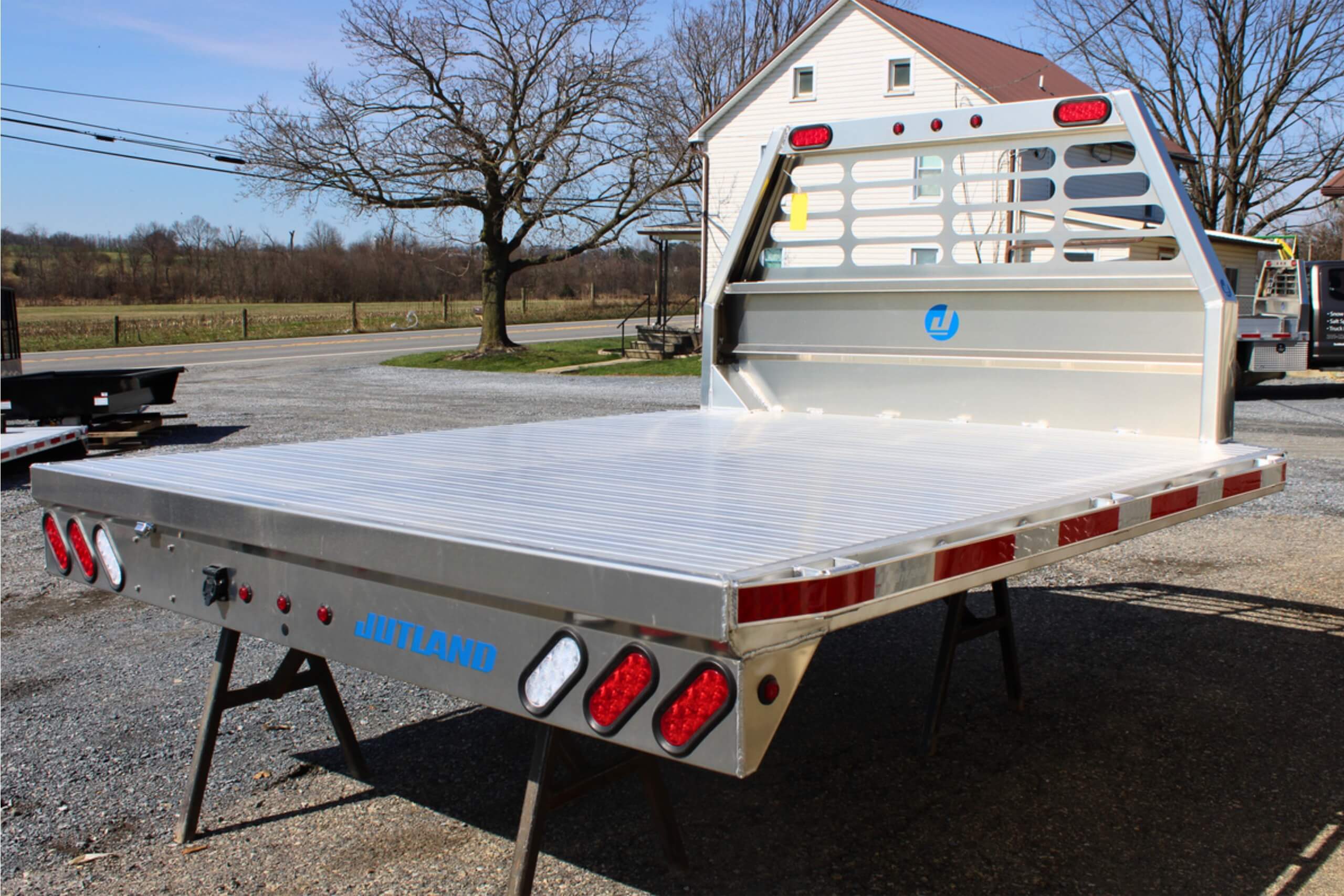 Aluminum flatbed truck body with red and white safety lights, reflective tape, and a headache rack, parked outdoors on gravel.
