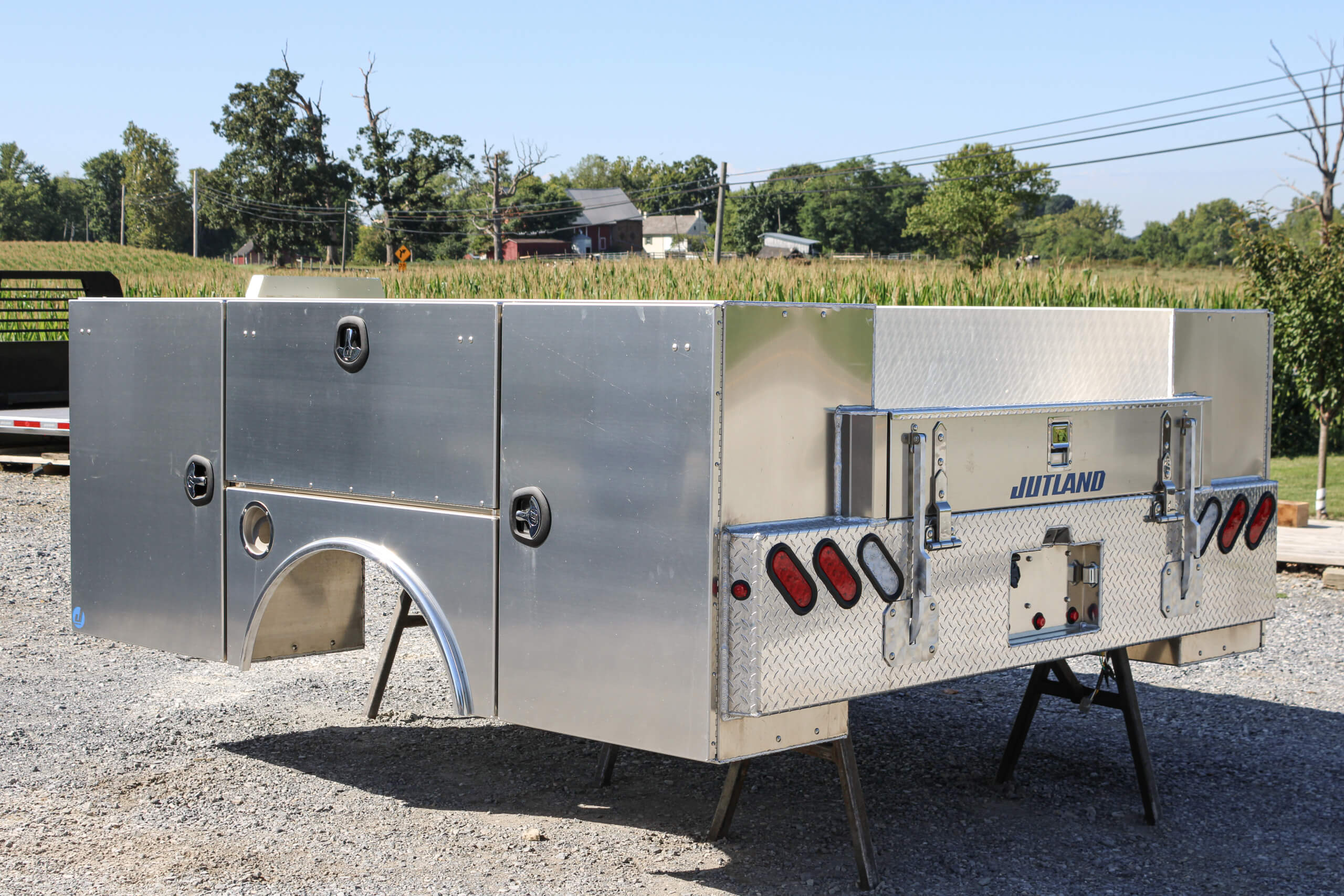 Aluminum truck utility bed with storage compartments and lights is elevated on two metal stands in an outdoor lot, with trees and buildings in the background.