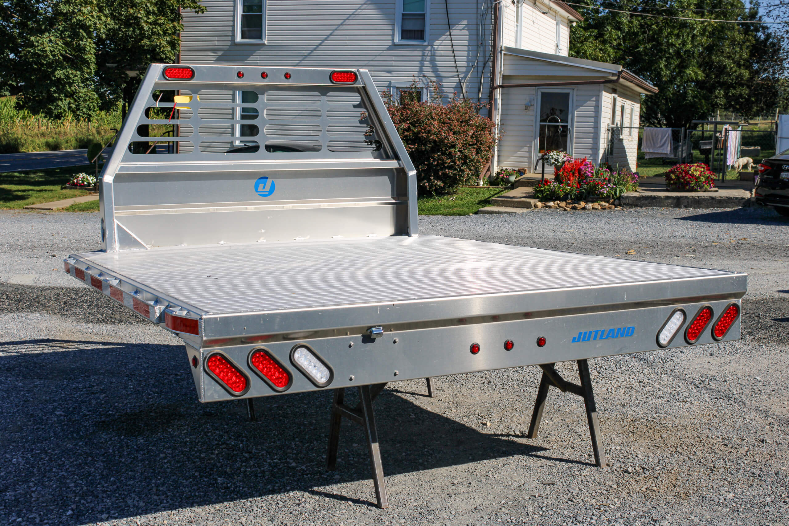 A silver aluminum flatbed truck body is displayed on metal stands outdoors, in front of a house and garden, under daylight.
