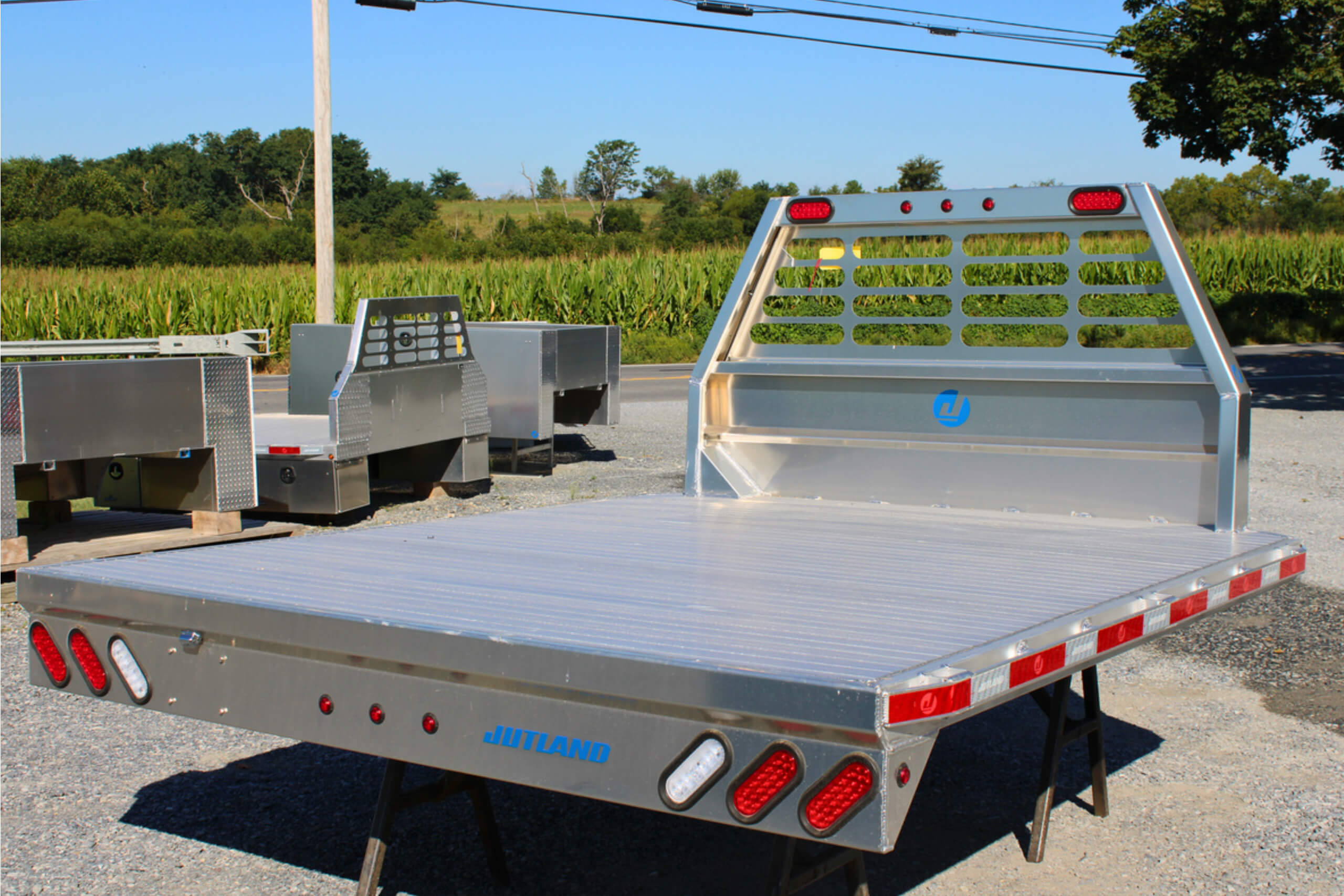 Aluminum flatbed truck body with red and white lights and reflective markings, displayed outdoors on stands with other truck beds in the background.