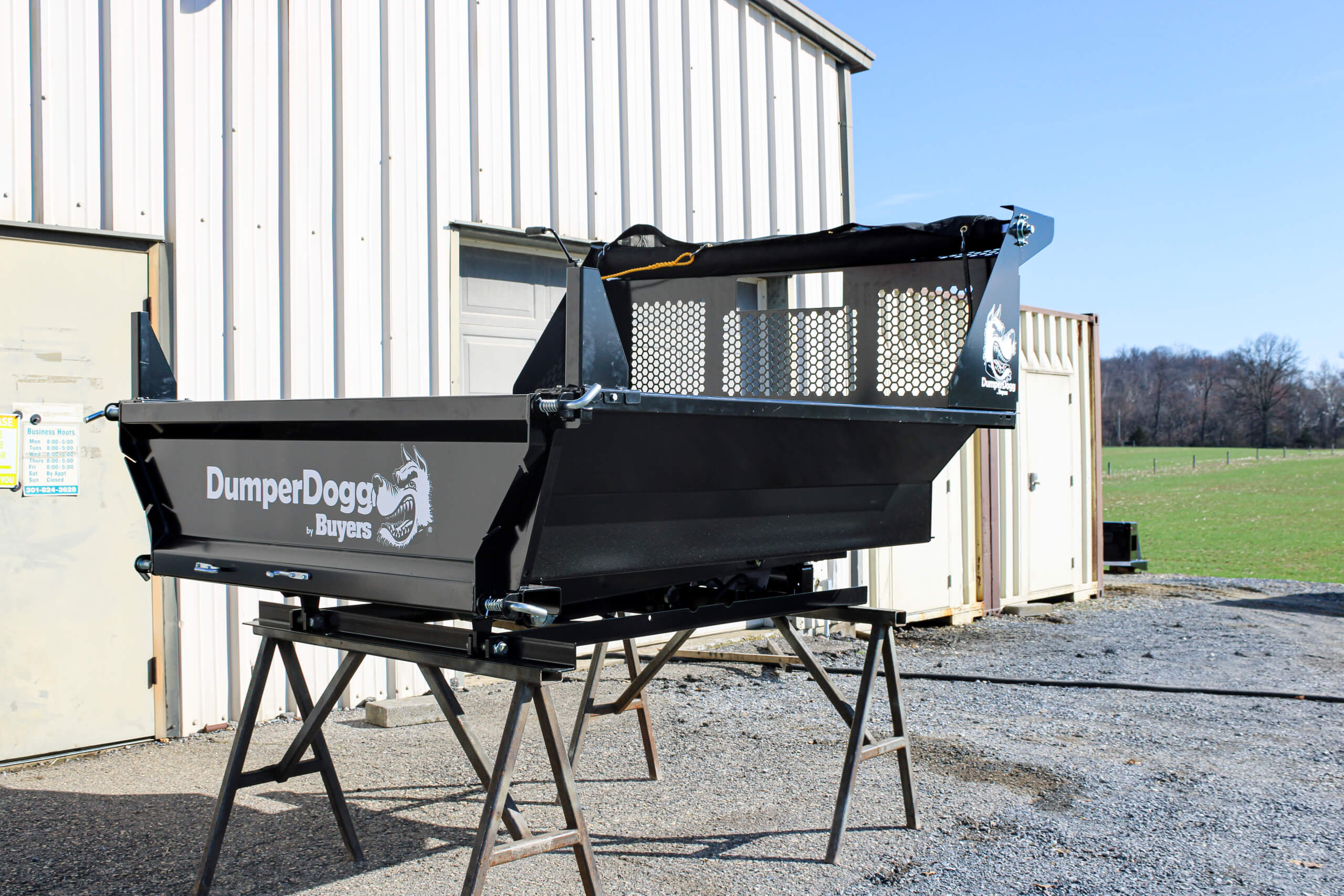 A black DumperDogg dump insert is displayed on a metal stand outside a light-colored metal building, with grass and trees in the background.