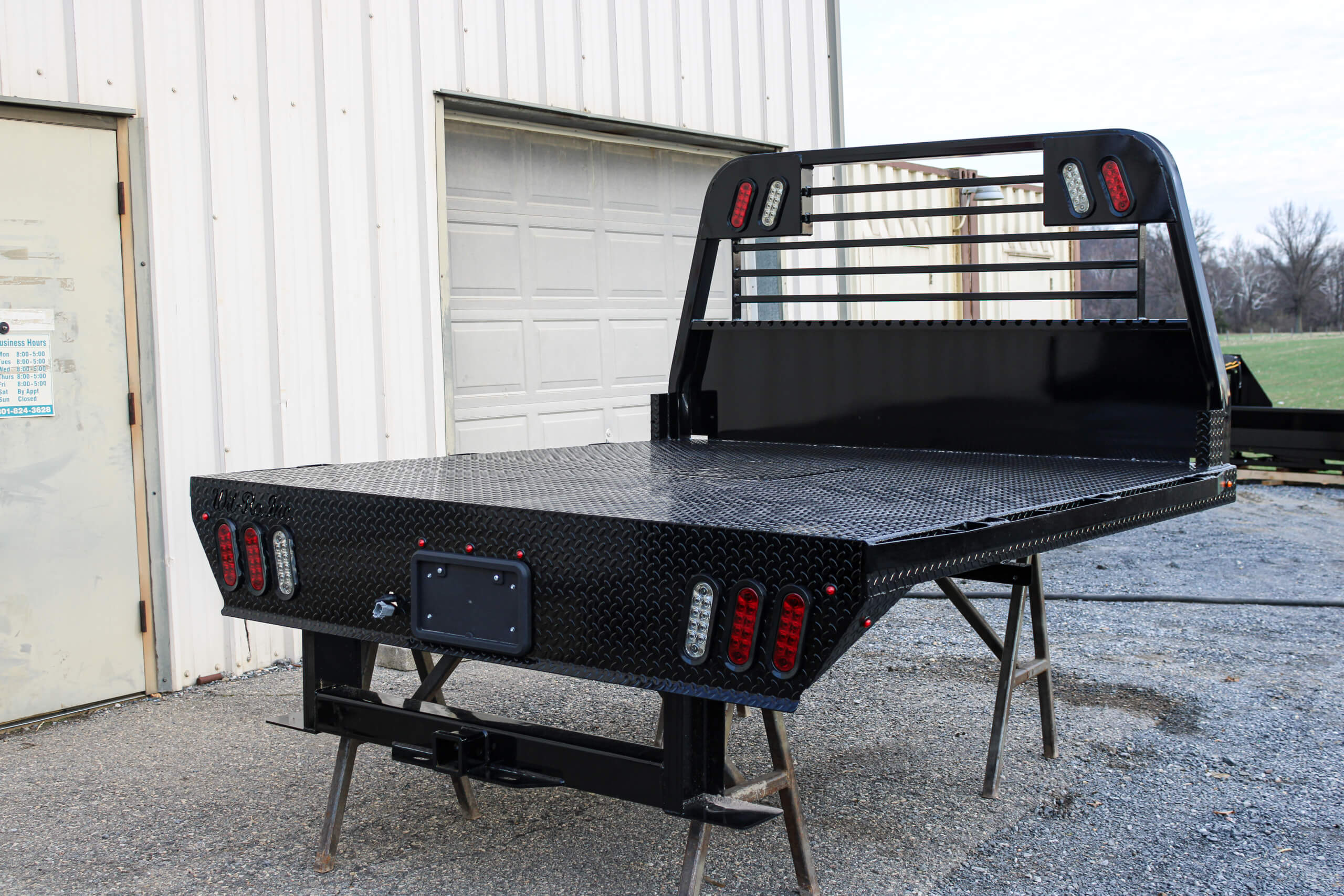 A black metal flatbed truck body with integrated lights is mounted on stands outside a metal building near a garage door.