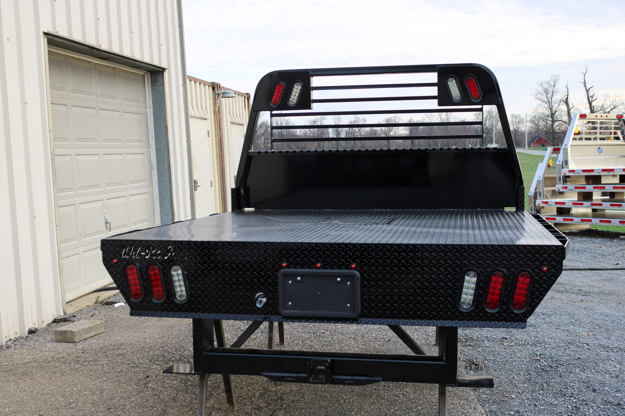 A black flatbed truck bed with lights is mounted on a frame outside a building near a closed garage door.