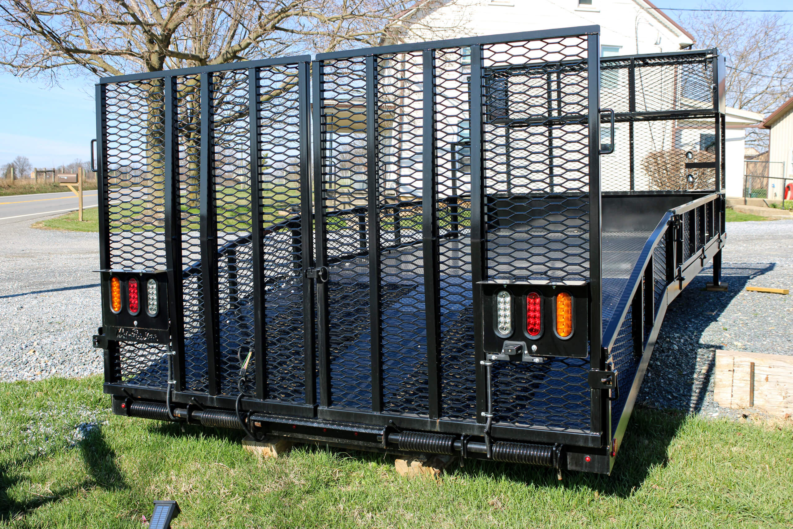 Black metal utility trailer with a mesh ramp gate is parked on grass, with red and amber tail lights visible. A house and trees are in the background.