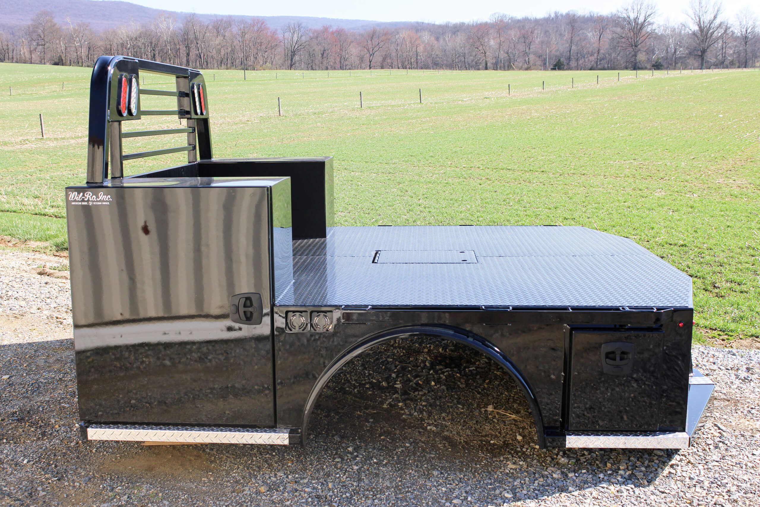 A black flatbed truck body with storage compartments sits on gravel near a grassy field and trees, with hills visible in the background.
