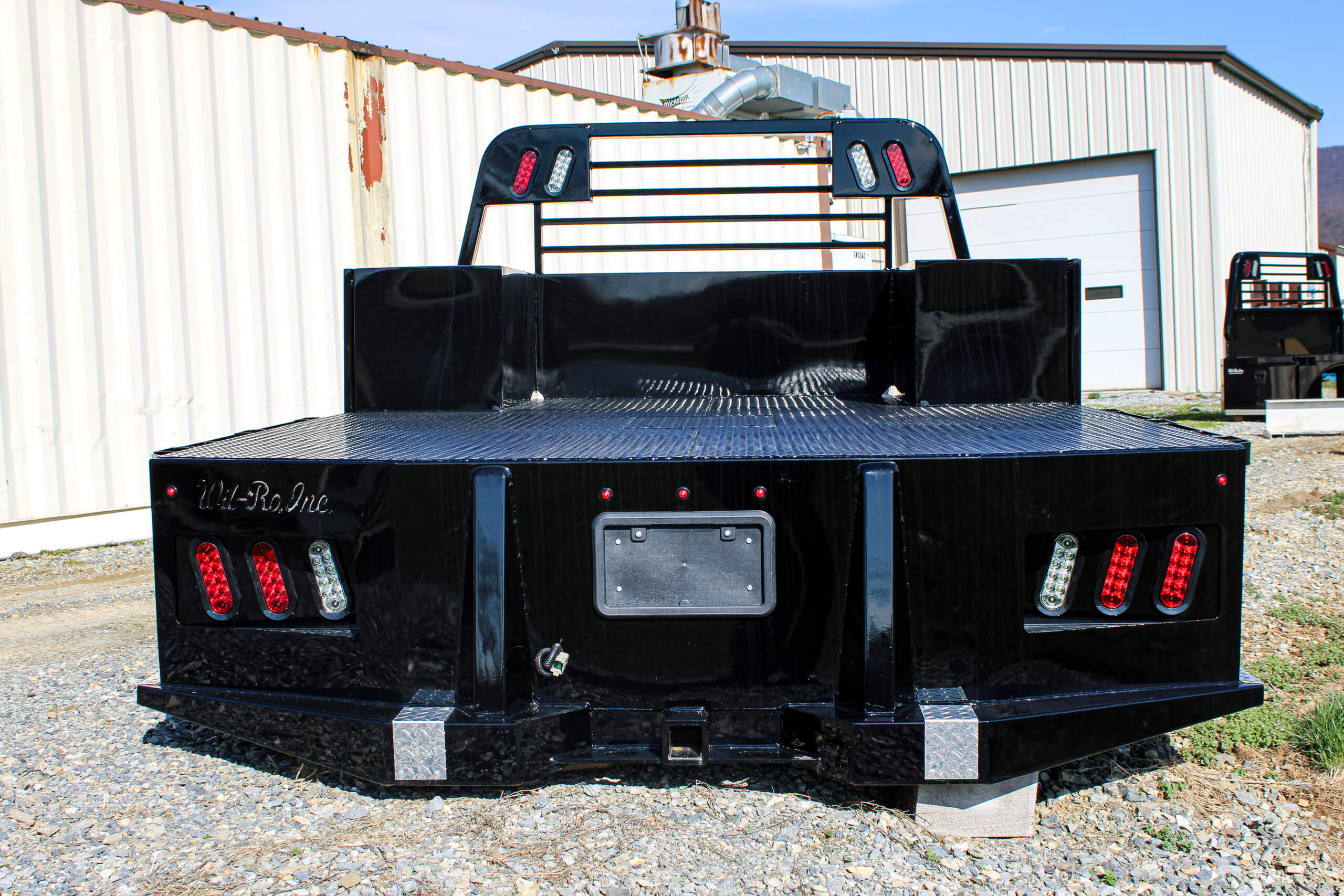 Black flatbed truck bed with red and white taillights, parked on gravel outside a metal building with a garage door in the background.