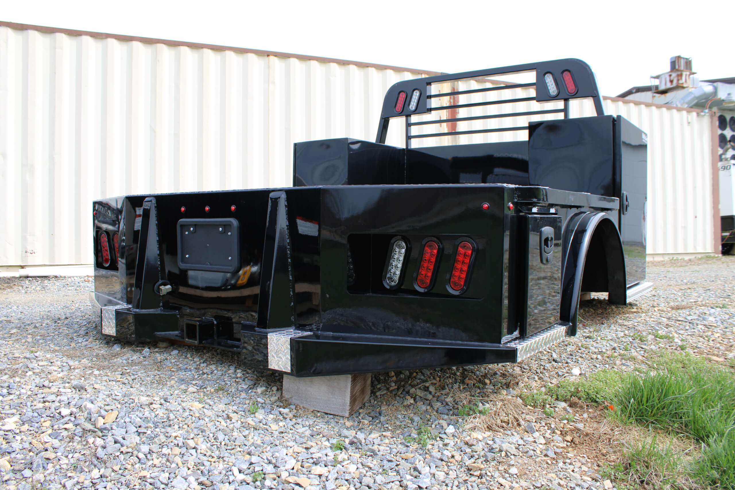 Black utility truck bed with toolboxes and rear lights, displayed outdoors on gravel, propped up by wooden blocks, with a shipping container in the background.