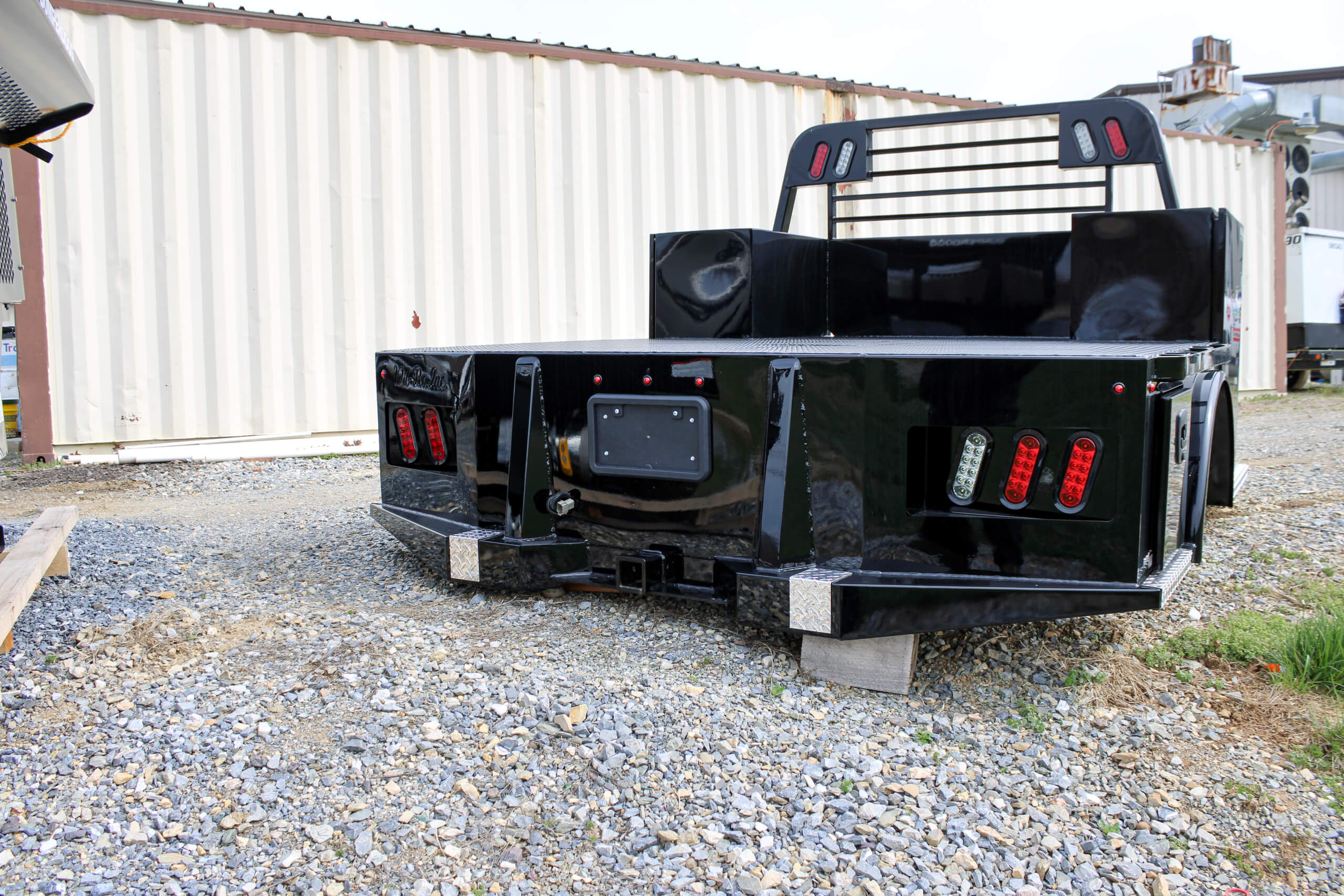 A black flatbed truck body sits on concrete blocks on gravel ground in front of a beige shipping container.