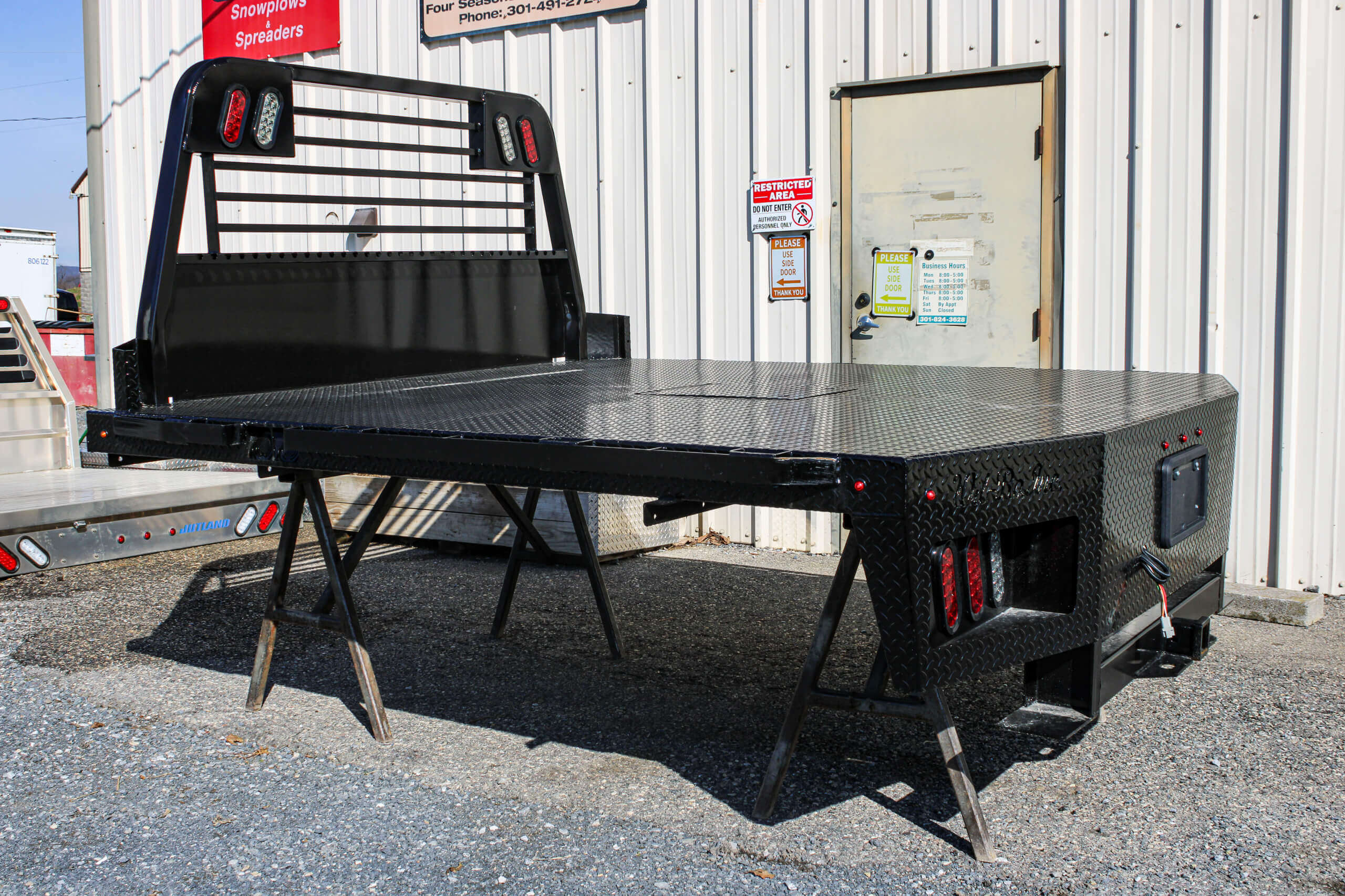 A black flatbed truck body is propped up on metal stands outside a metal building, near a closed door with warning signs.