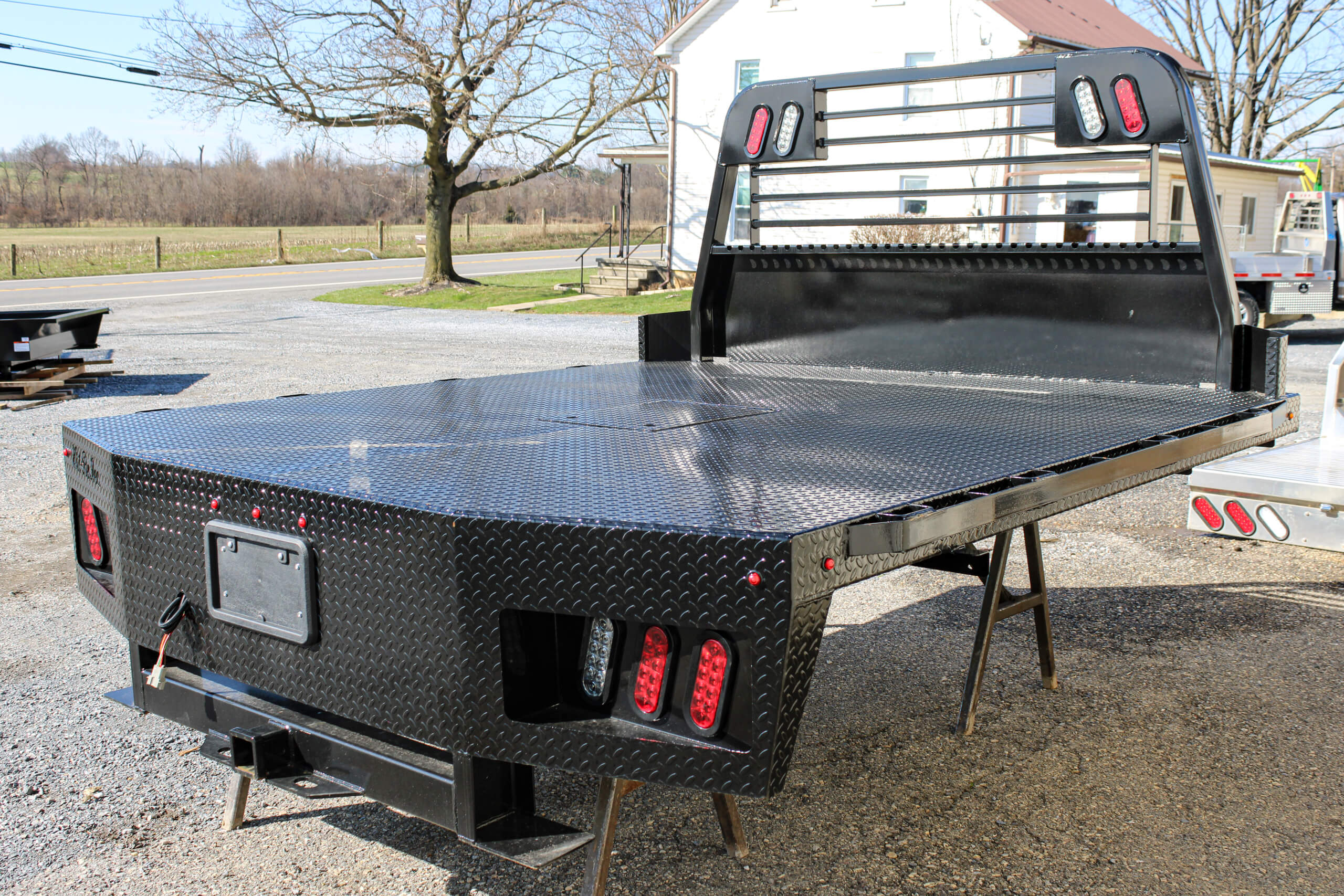 A black diamond plate steel flatbed truck bed with built-in red tail lights is set on stands outdoors near a house and an open field.