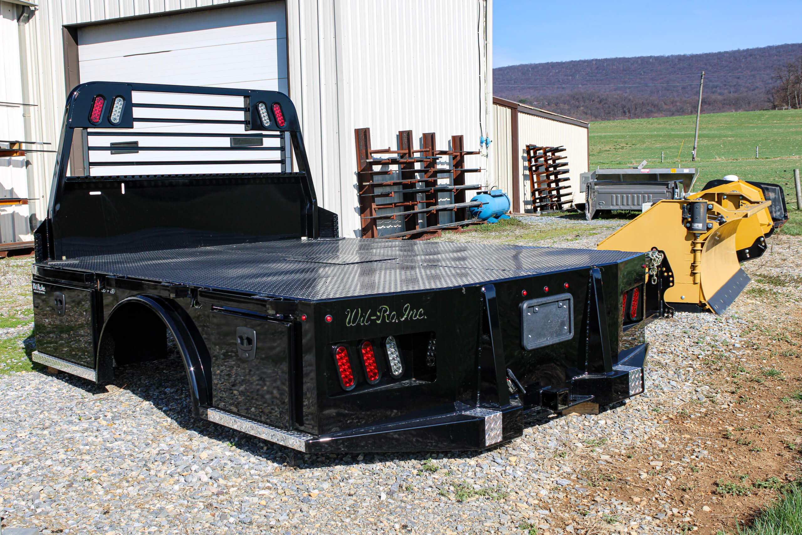 A black Wil-Ro flatbed truck body is parked on gravel outside a metal building, with farm equipment and a grassy field in the background.