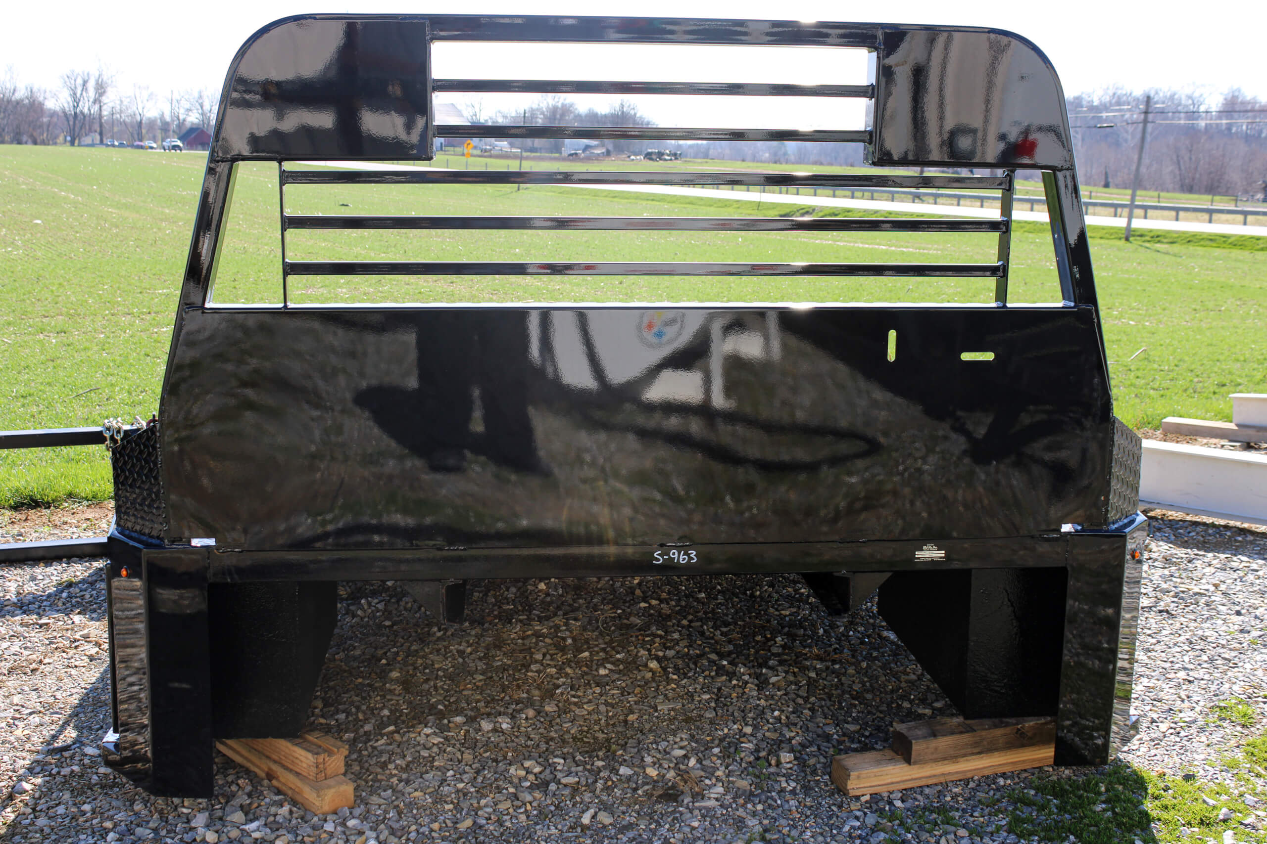 A black metal truck bed with a headache rack is displayed outdoors on gravel, supported by wooden blocks, with a grassy field and road in the background.