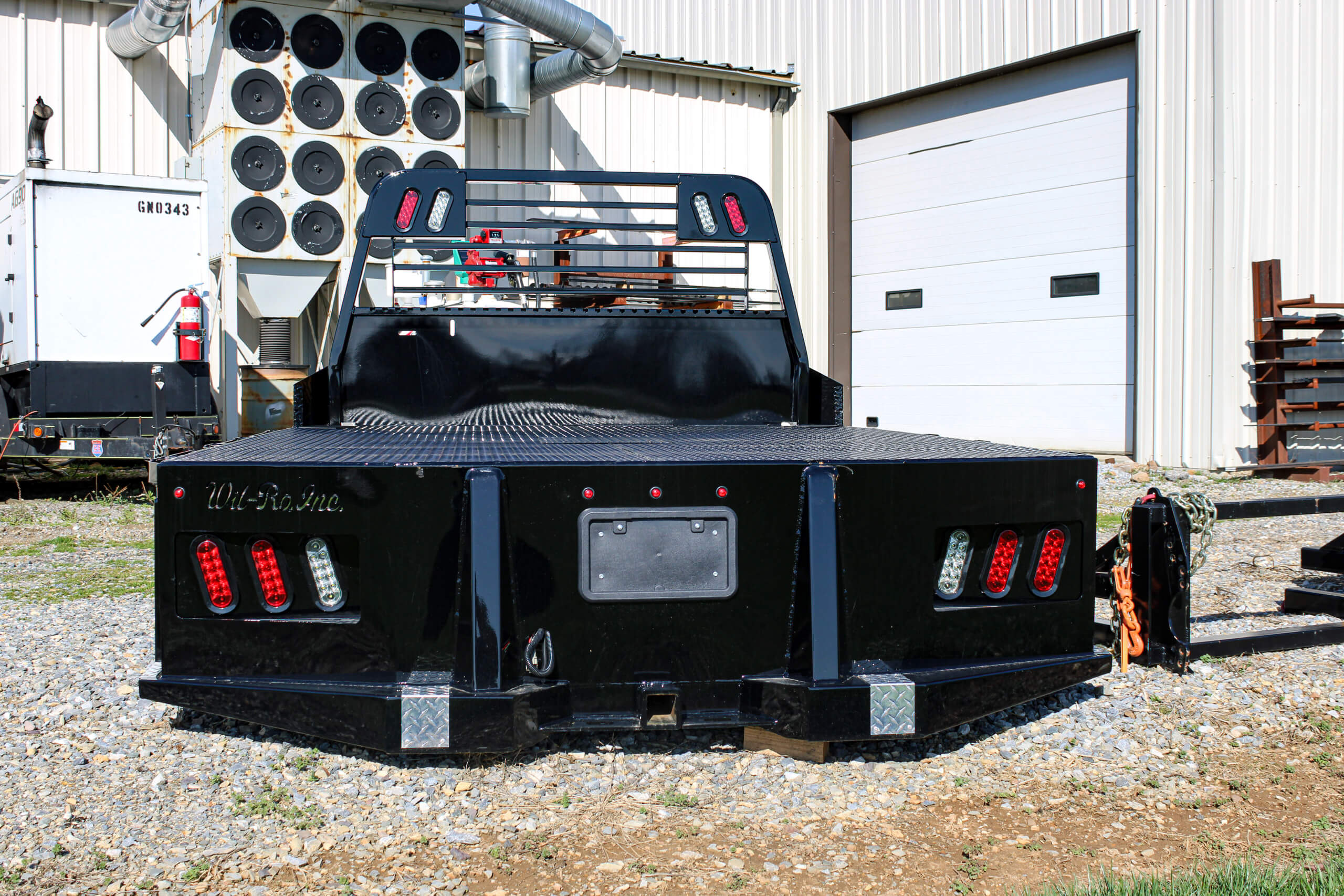 A black flatbed truck bed with red and white lights is parked on gravel in front of a metal building and garage door.