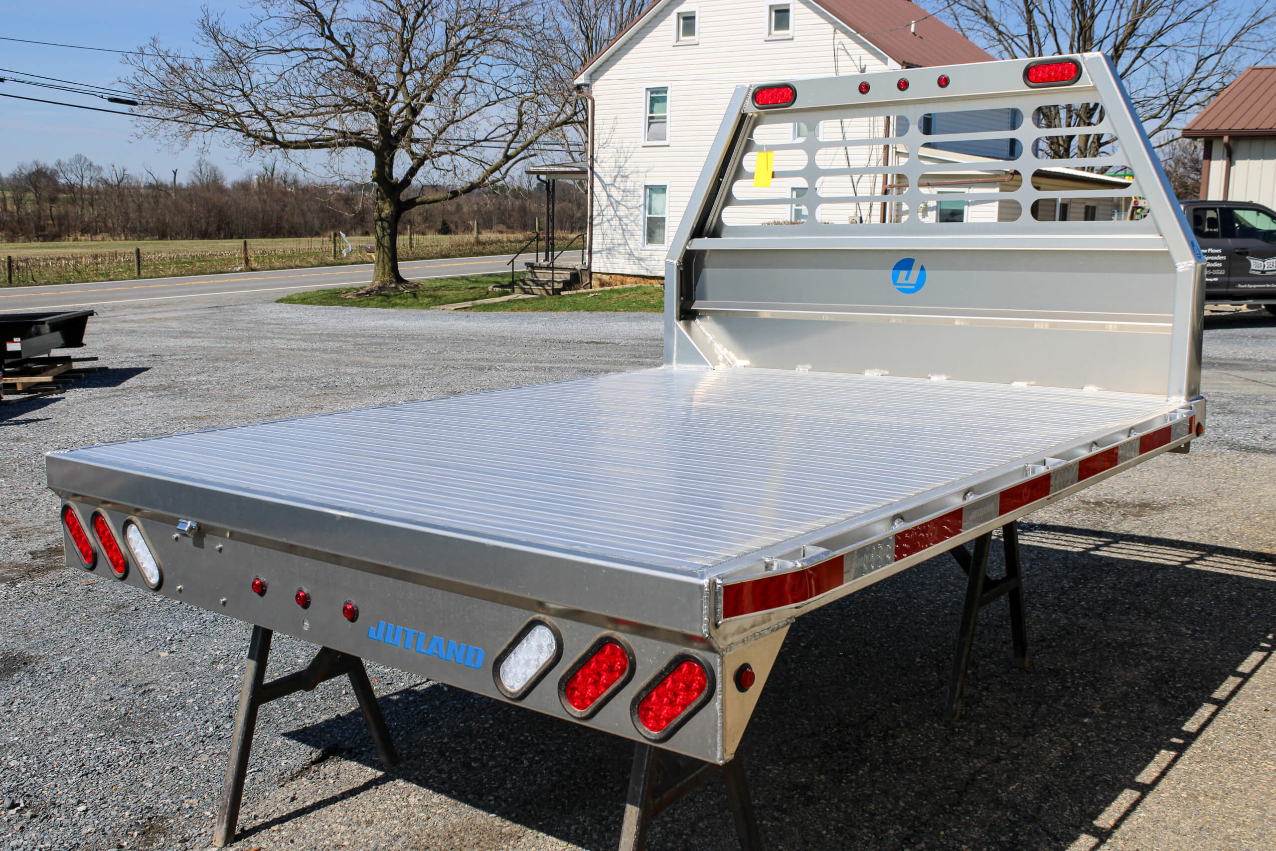 Aluminum flatbed truck body with red and white lights, mounted on stands in an outdoor lot near a white house and trees.