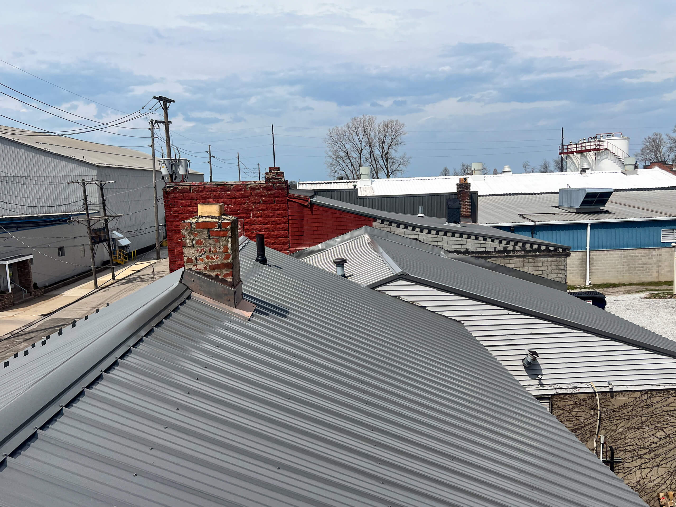 Crawford County Roofing Spartansburg, PA Rooftops with metal sheets, brick chimneys, and utility poles under a partly cloudy sky in an industrial area.