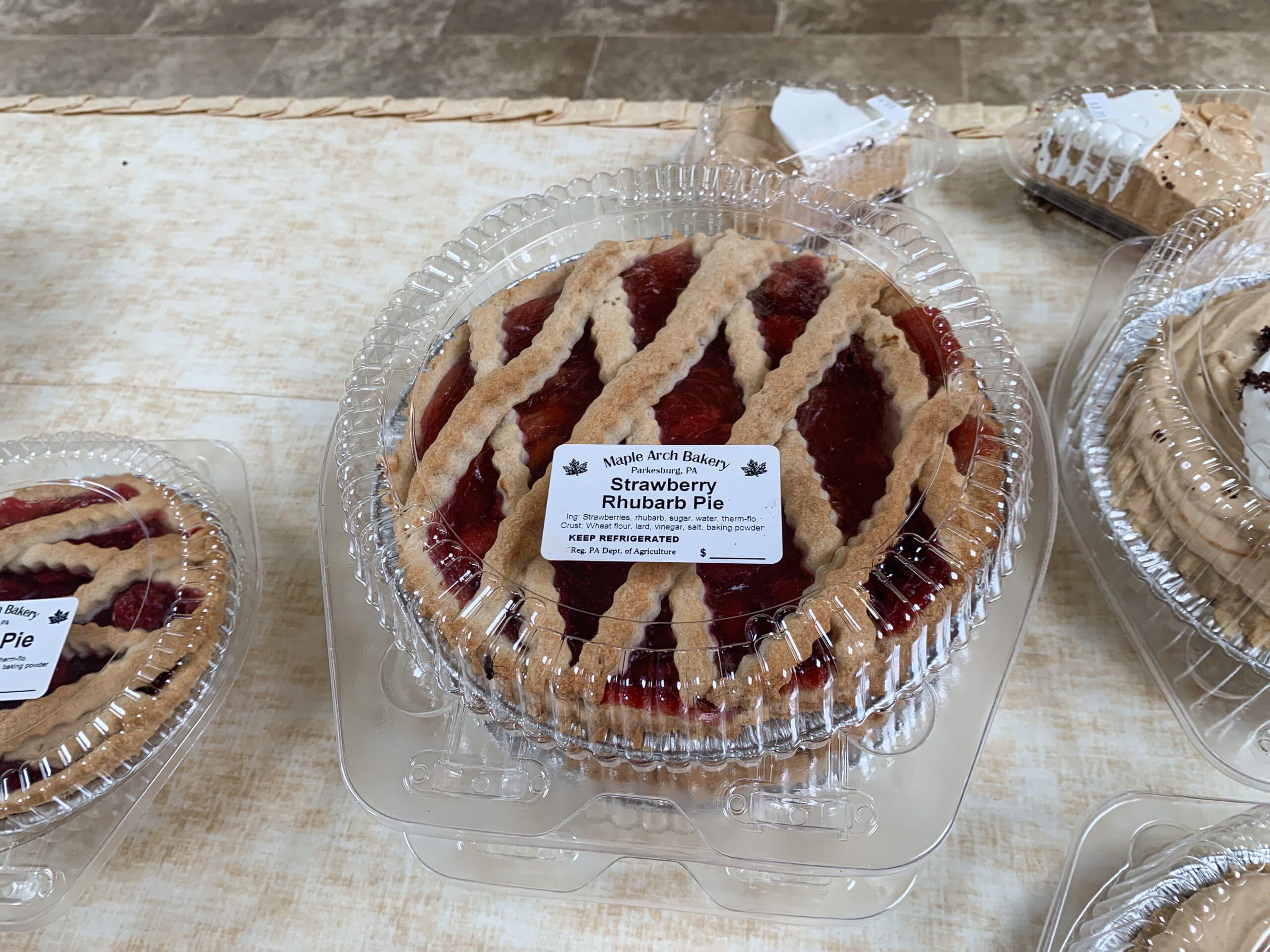 Maple Arch Farm Market Cochranville PA A strawberry rhubarb pie with a lattice crust is packaged in a clear plastic container and labeled from Maple Arch Bakery, displayed on a table with other pies.