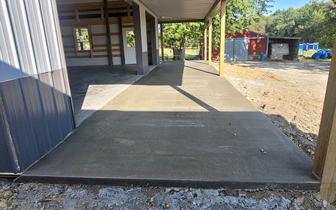 J&S Concrete Hawesville KY Freshly poured concrete sidewalk under a metal roof structure beside a partially constructed building, with construction materials and debris visible in the background.