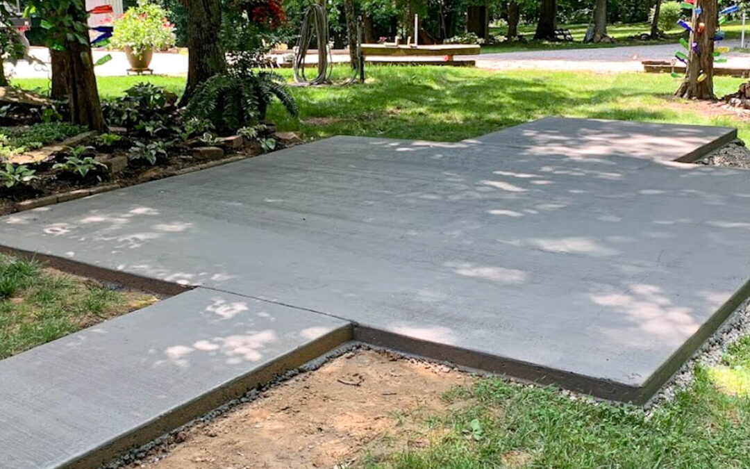J&S Concrete Hawesville KY Freshly poured and smoothed concrete slab with a short walkway in a shaded, grassy outdoor area surrounded by trees and plants.