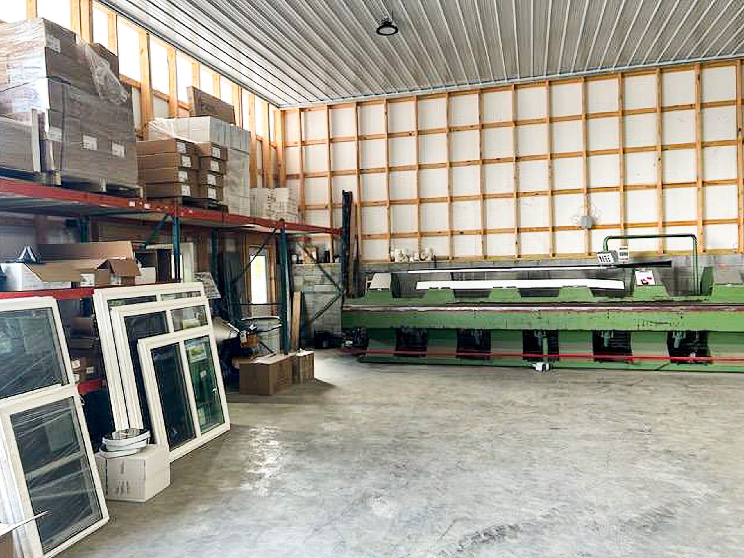 Metal Profiles Rose Hill, VA A warehouse interior with shelves of boxes, stacked window frames, and a large green industrial machine against a wall with exposed wooden framing.