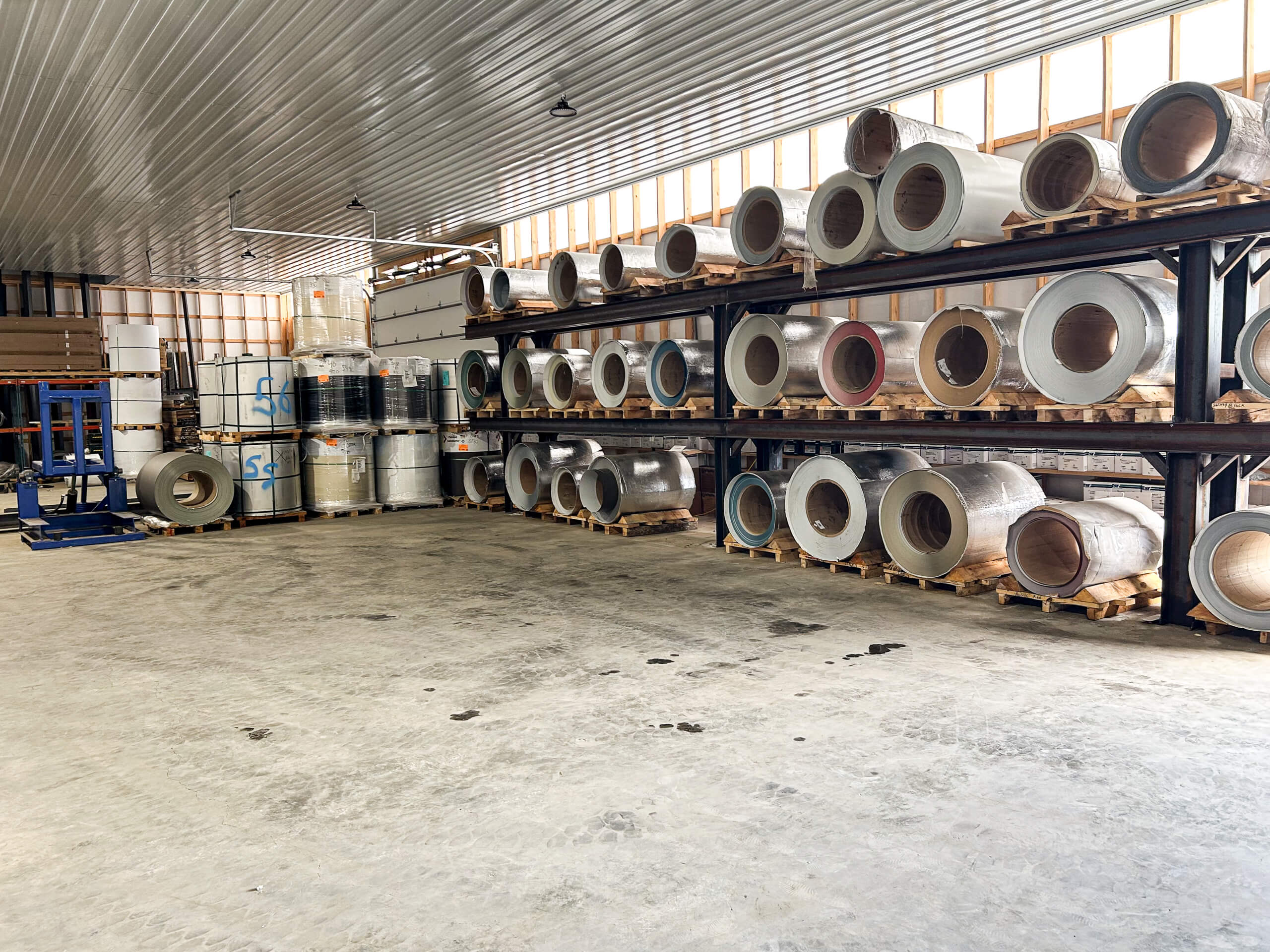 Metal Profiles Rose Hill, VA Large warehouse interior with metal coils stored on heavy-duty shelves along the wall; stacks of barrels and equipment are visible on the left side of the space.