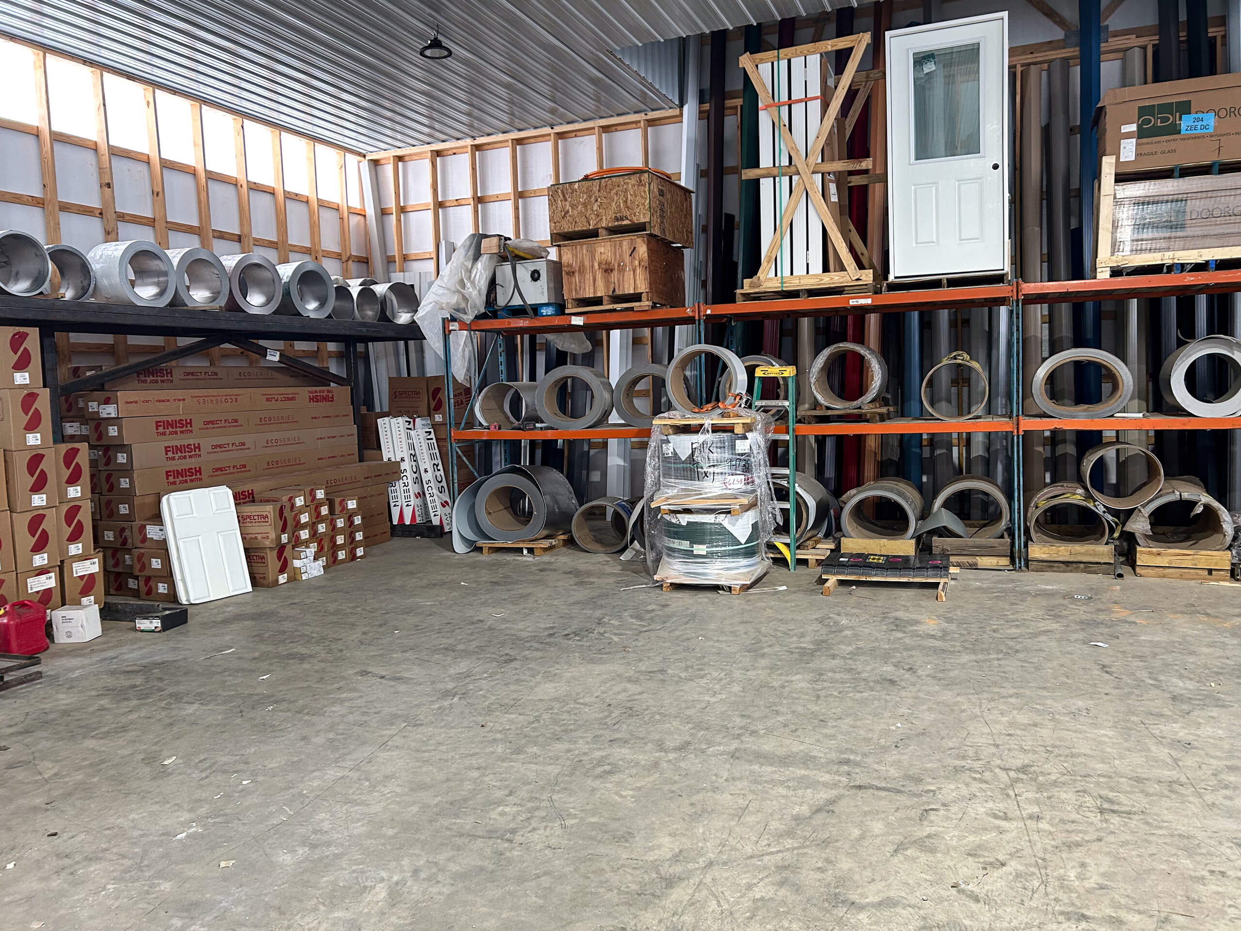 Metal Profiles Rose Hill, VA Industrial warehouse interior with metal coils, construction materials, stacked boxes, and a door stored on shelves against the wall. The floor is mostly clear.